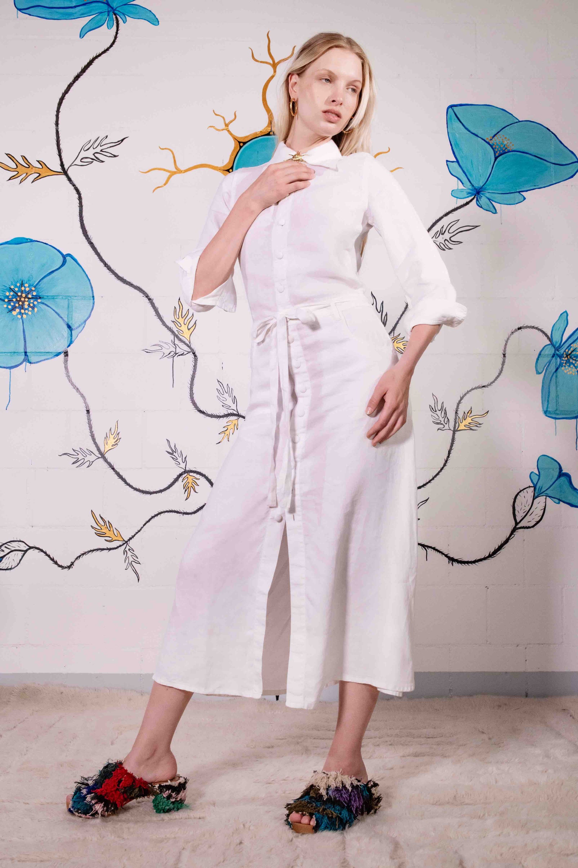 Model is wearing the Blanc Cassé Trompe L’Oeil dress by Happy Haus, made from a soft hemp and silk blend with a classic collar, bronze buttons, 7/8-length pleated sleeves, and floral patch pockets including peacock embroidery.