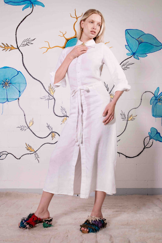 Model is wearing the Blanc Cassé Trompe L’Oeil dress by Happy Haus, made from a soft hemp and silk blend with a classic collar, bronze buttons, 7/8-length pleated sleeves, and floral patch pockets including peacock embroidery.