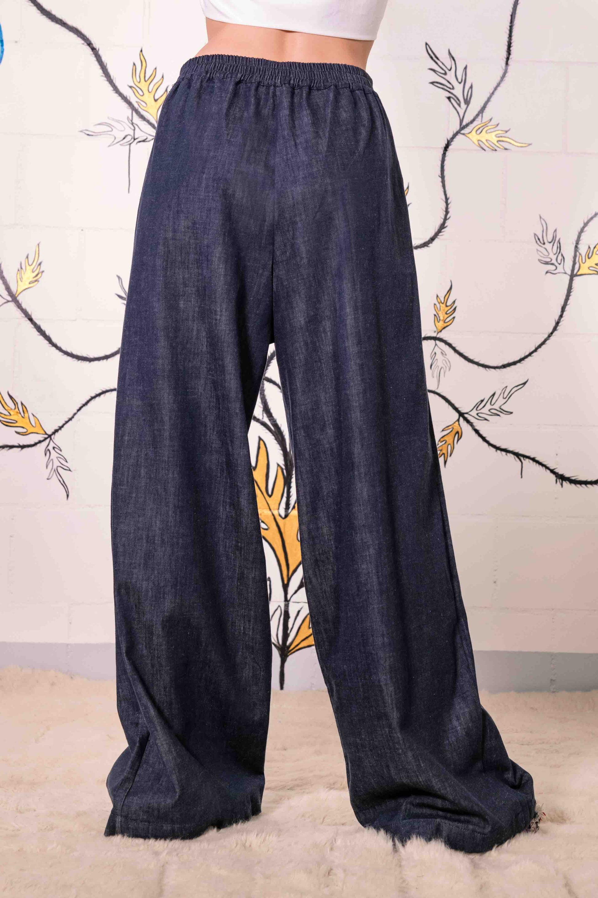 Model is wearing dark denim wide-leg trousers made from 100% Italian cotton, featuring a mid-rise elastic waistband, front pleats, and side pockets.