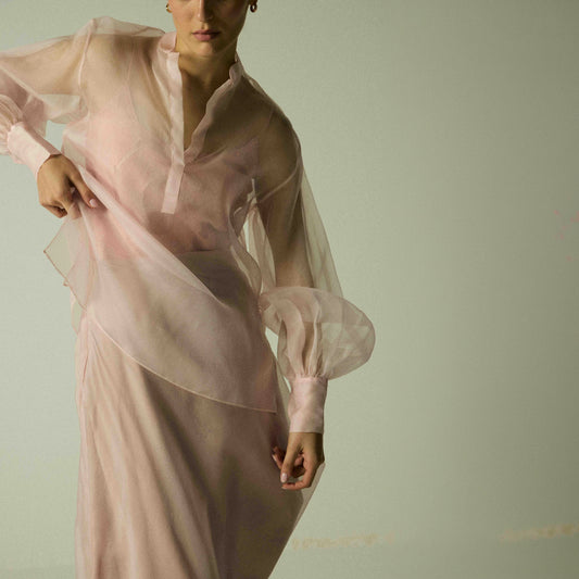 Model wearing a pink silk organza shirt by Abadia from the House of Nomad Stories collection, featuring sheer balloon sleeves and a lightweight structured silhouette in an elegant minimal setting.