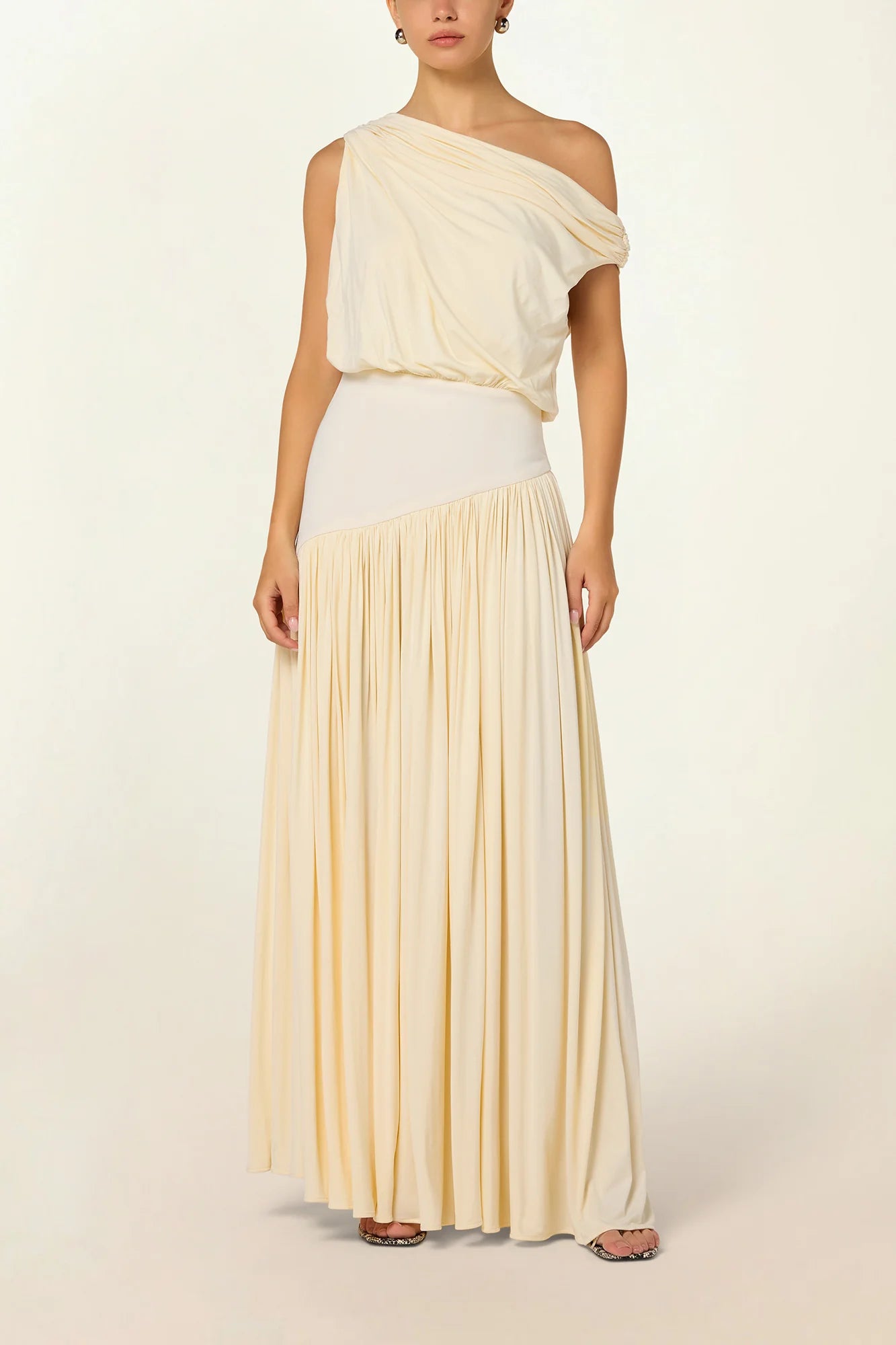 Model wearing a cream Gwen Off Shoulder Drape Dress by Abadia from the House of Nomad Stories collection, featuring a stretch jersey fabric, asymmetrical curved waistband, and a sculpted elegant maxi silhouette.