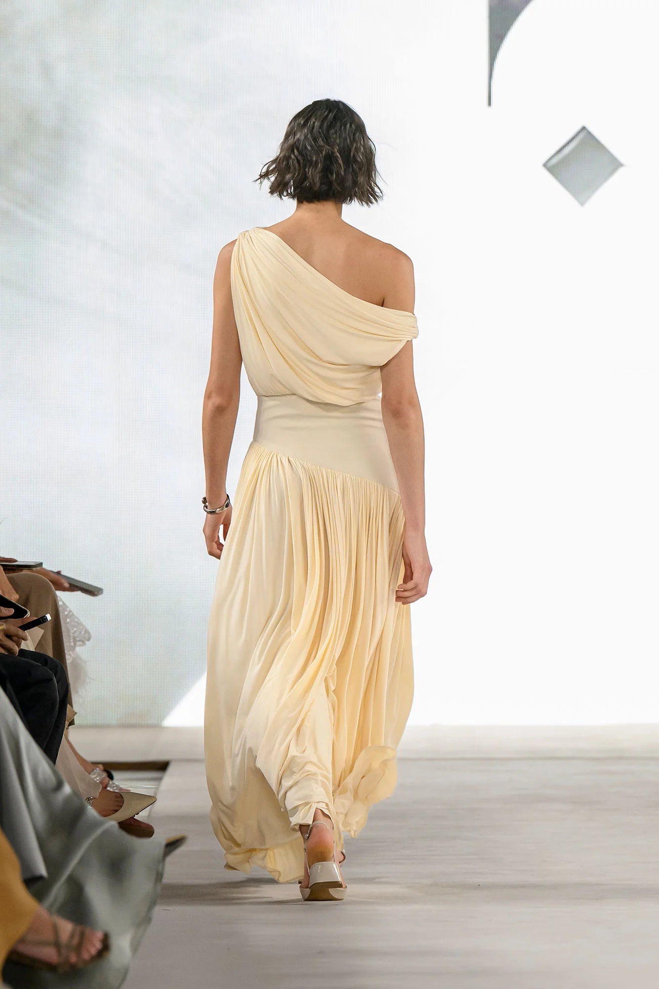Model wearing a cream Gwen Off Shoulder Drape Dress by Abadia from the House of Nomad Stories collection, featuring a stretch jersey fabric, asymmetrical curved waistband, and a sculpted elegant maxi silhouette.