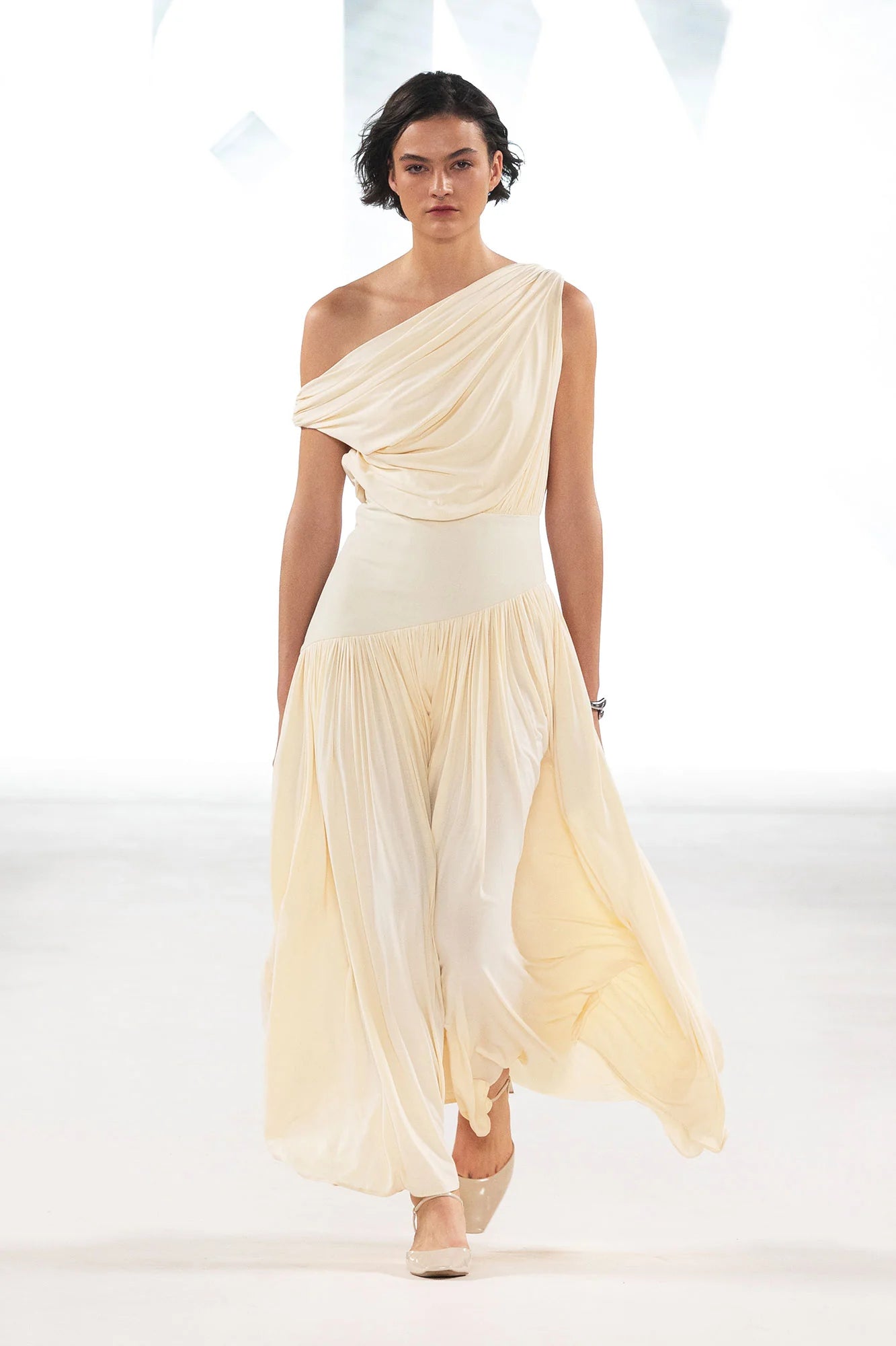 Model wearing a cream Gwen Off Shoulder Drape Dress by Abadia from the House of Nomad Stories collection, featuring a stretch jersey fabric, asymmetrical curved waistband, and a sculpted elegant maxi silhouette.