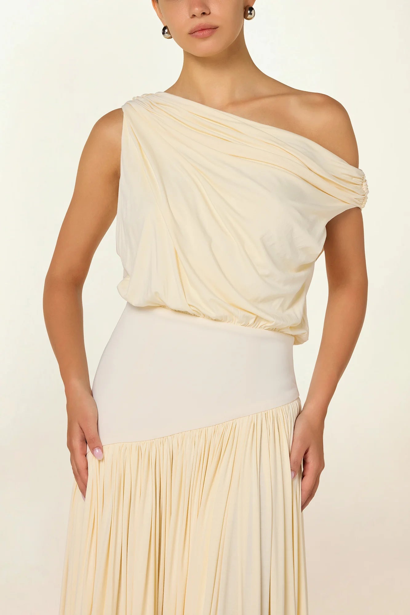 Model wearing a cream Gwen Off Shoulder Drape Dress by Abadia from the House of Nomad Stories collection, featuring a stretch jersey fabric, asymmetrical curved waistband, and a sculpted elegant maxi silhouette.