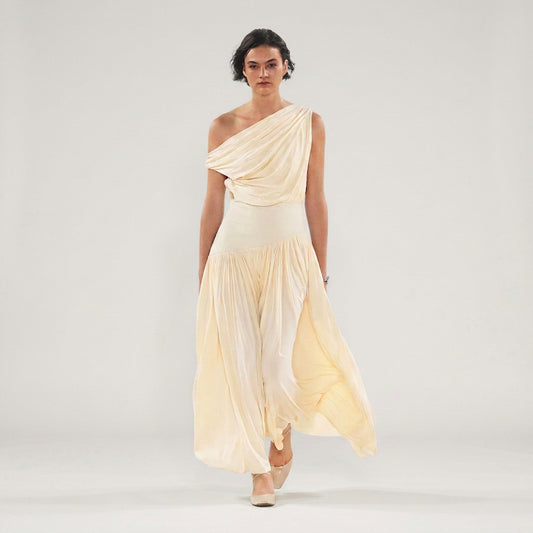 Model wearing a cream Gwen Off Shoulder Drape Dress by Abadia from the House of Nomad Stories collection, featuring a stretch jersey fabric, asymmetrical curved waistband, and a sculpted elegant maxi silhouette.