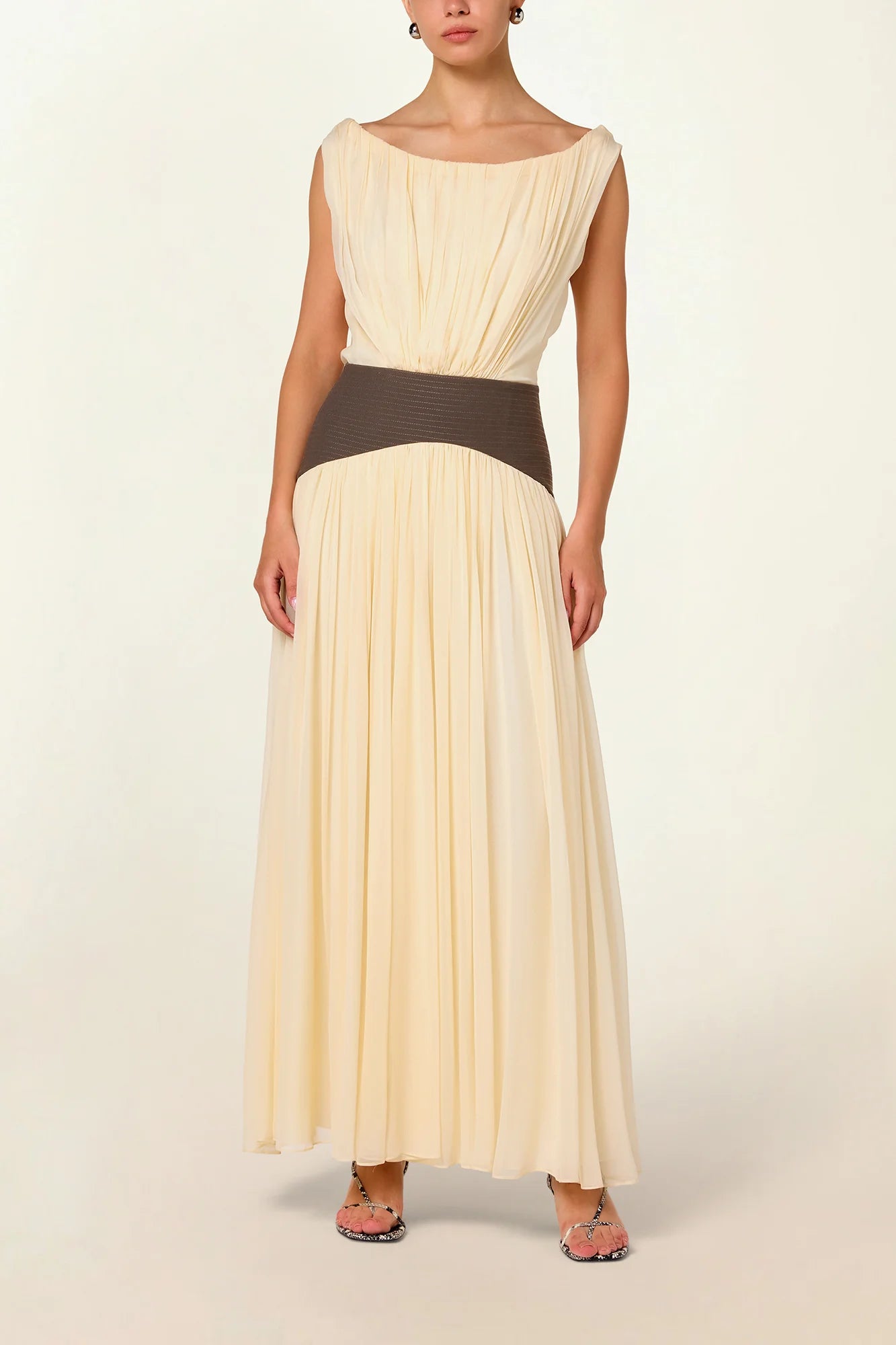 Model wearing a cream Siena Maxi Dress by Abadia from the House of Nomad Stories collection, featuring an off-shoulder draped bodice, sculpted green waistband, and a flowing elegant maxi silhouette.