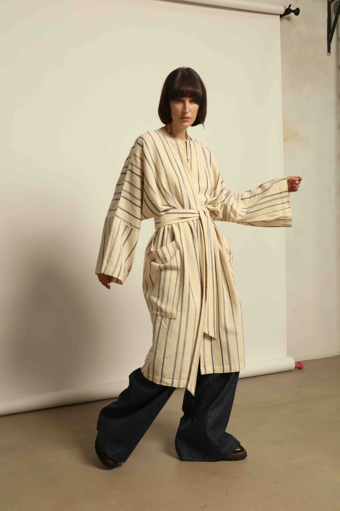 Model is wearing the Maxi Handwoven and Hand-Printed Benin Cotton Kimono ‘The Ones Who Laugh’ by Atelier V.O, crafted from 100% handwoven cotton with Dogon Mali-inspired print, straight cut, drawstring, wooden button fastening, and matching belt.