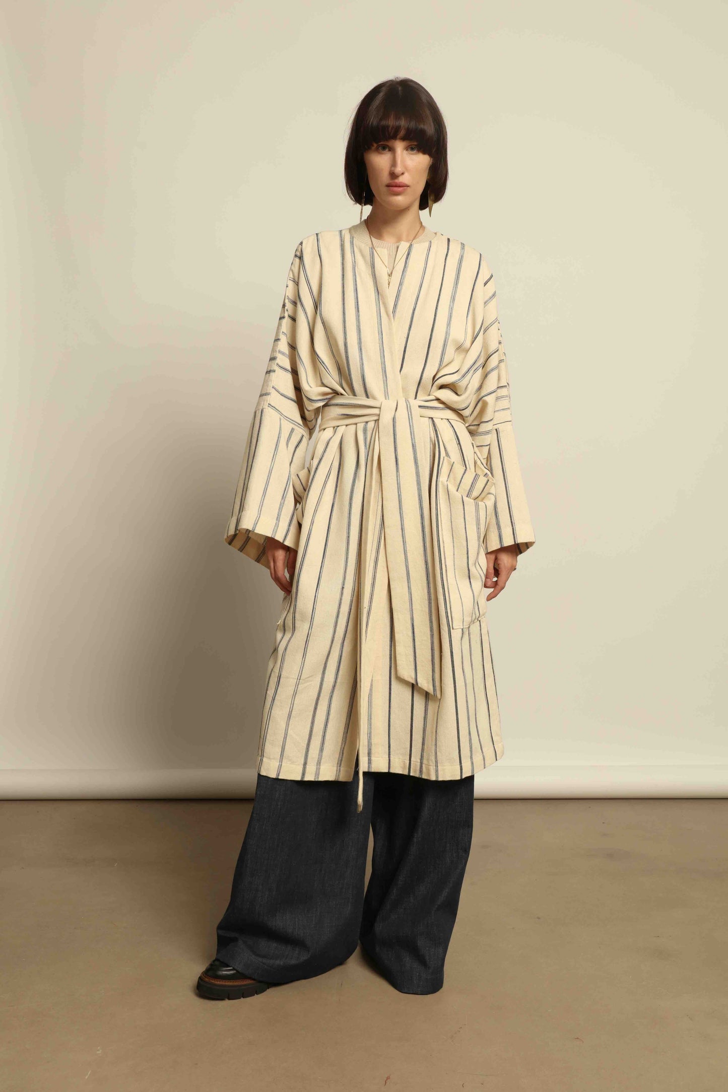 Model is wearing the Maxi Handwoven and Hand-Printed Benin Cotton Kimono ‘The Ones Who Laugh’ by Atelier V.O, crafted from 100% handwoven cotton with Dogon Mali-inspired print, straight cut, drawstring, wooden button fastening, and matching belt.