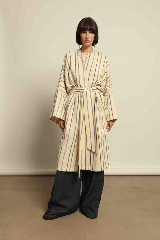 Model is wearing the Maxi Handwoven and Hand-Printed Benin Cotton Kimono ‘The Ones Who Laugh’ by Atelier V.O, crafted from 100% handwoven cotton with Dogon Mali-inspired print, straight cut, drawstring, wooden button fastening, and matching belt.
