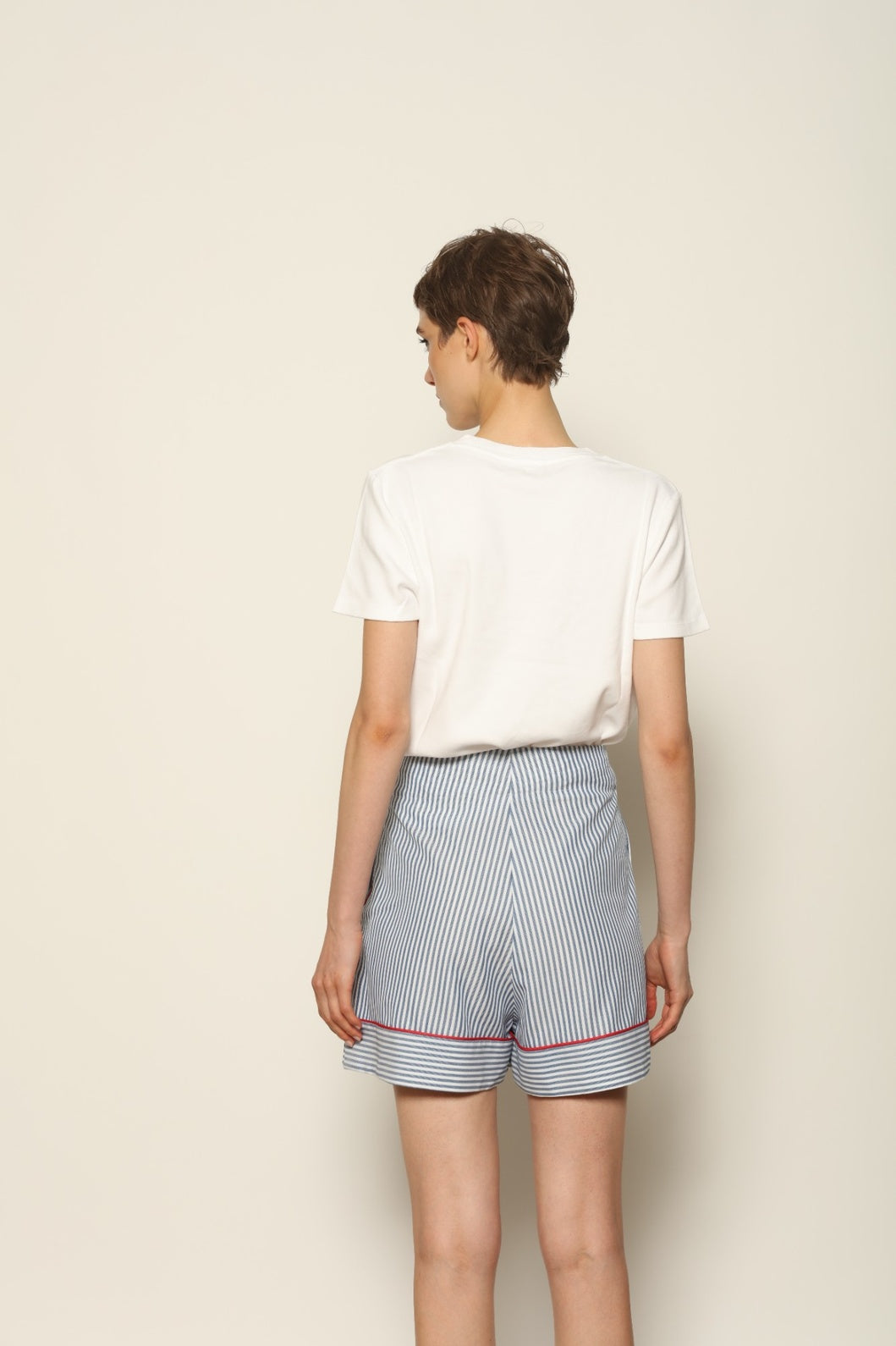 Model is wearing blue and white striped 100% cotton shorts with contrasting red piping by House of Woven Stories. The shorts feature a relaxed, tailored silhouette designed for comfort and ease of movement. Made in Italy from premium natural cotton, they reflect artisan craftsmanship and timeless warm-weather style, styled as part of a contemporary resort wear look.