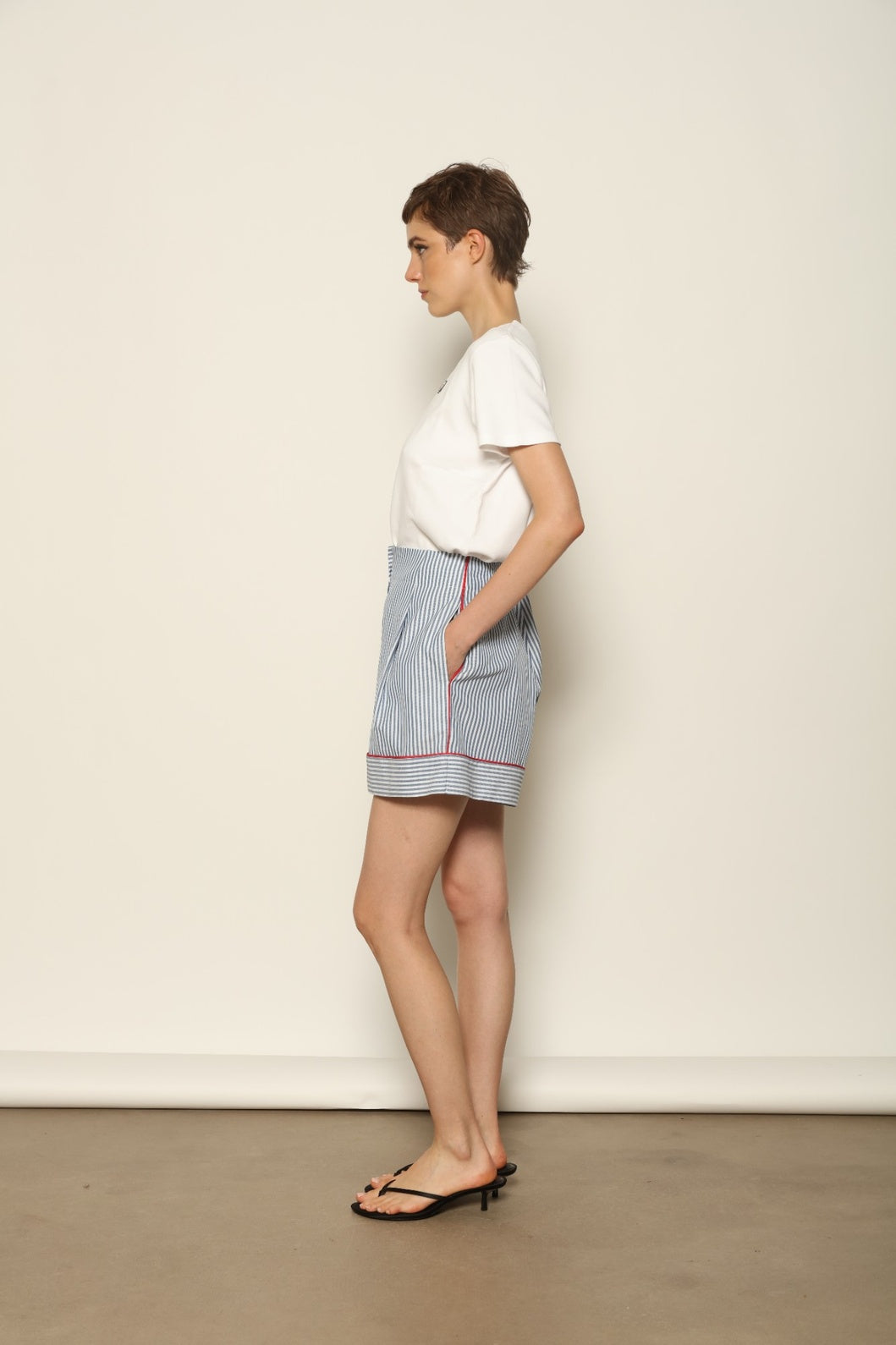 Model is wearing blue and white striped 100% cotton shorts with contrasting red piping by House of Woven Stories. The shorts feature a relaxed, tailored silhouette designed for comfort and ease of movement. Made in Italy from premium natural cotton, they reflect artisan craftsmanship and timeless warm-weather style, styled as part of a contemporary resort wear look.