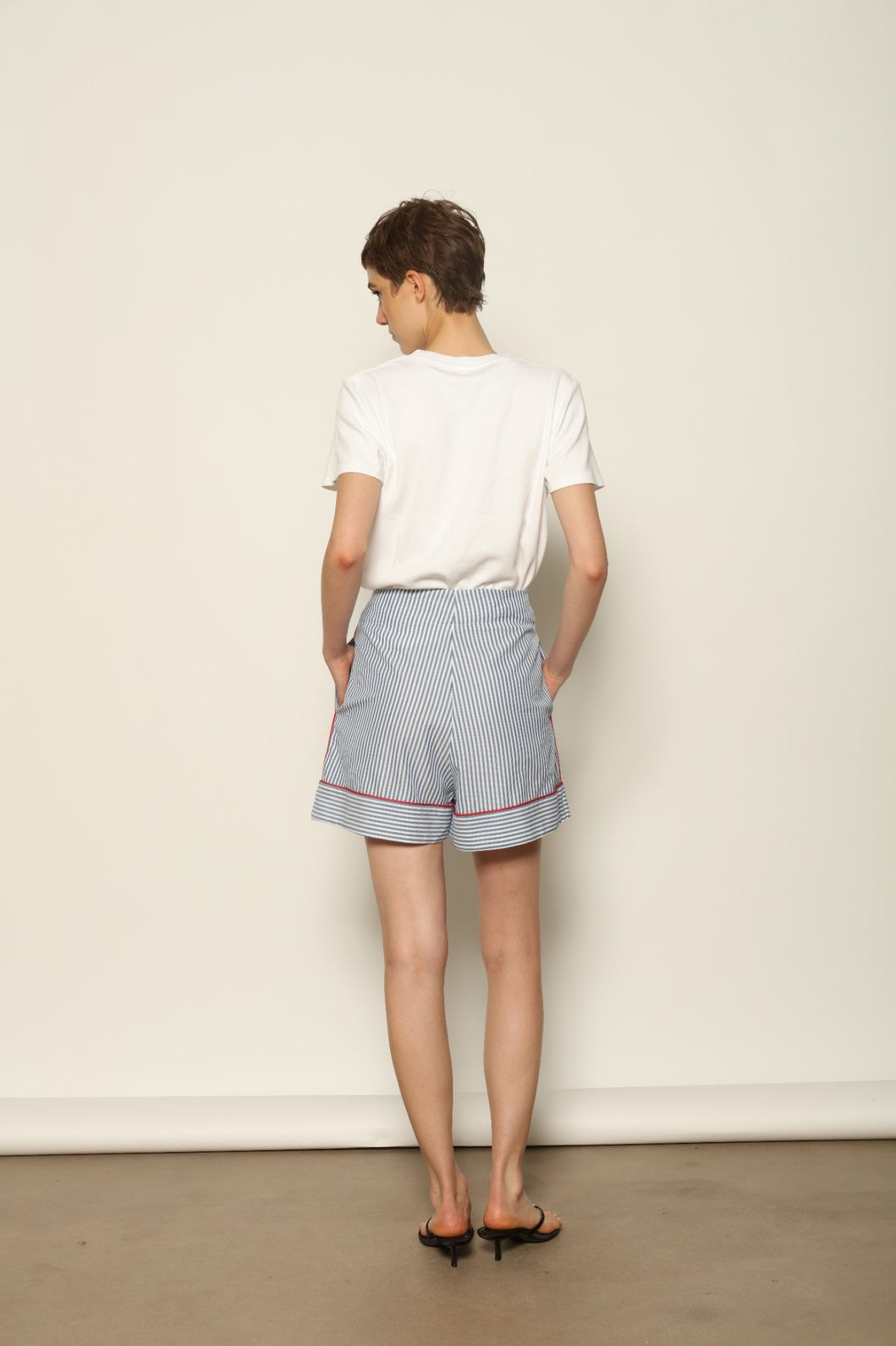 Model is wearing blue and white striped 100% cotton shorts with contrasting red piping by House of Woven Stories. The shorts feature a relaxed, tailored silhouette designed for comfort and ease of movement. Made in Italy from premium natural cotton, they reflect artisan craftsmanship and timeless warm-weather style, styled as part of a contemporary resort wear look.