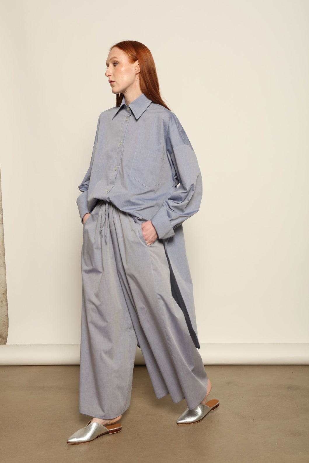 Model is wearing the Brigitta dusty blue cotton mousseline trousers from the House of Woven Stories selection, lightweight and breathable women’s summer pants made in Italy in Piedmont, crafted from 100% natural cotton with a relaxed tailored silhouette designed for effortless movement.
