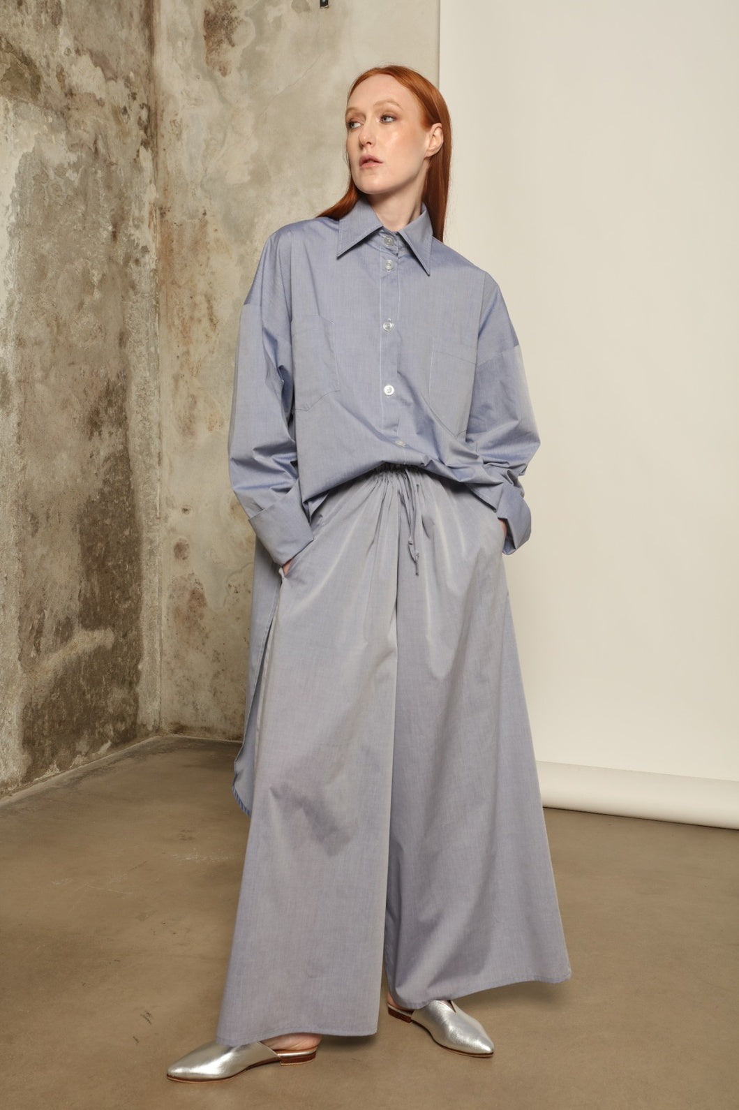 Model is wearing the Brigitta dusty blue cotton mousseline trousers from the House of Woven Stories selection, lightweight and breathable women’s summer pants made in Italy in Piedmont, crafted from 100% natural cotton with a relaxed tailored silhouette designed for effortless movement.