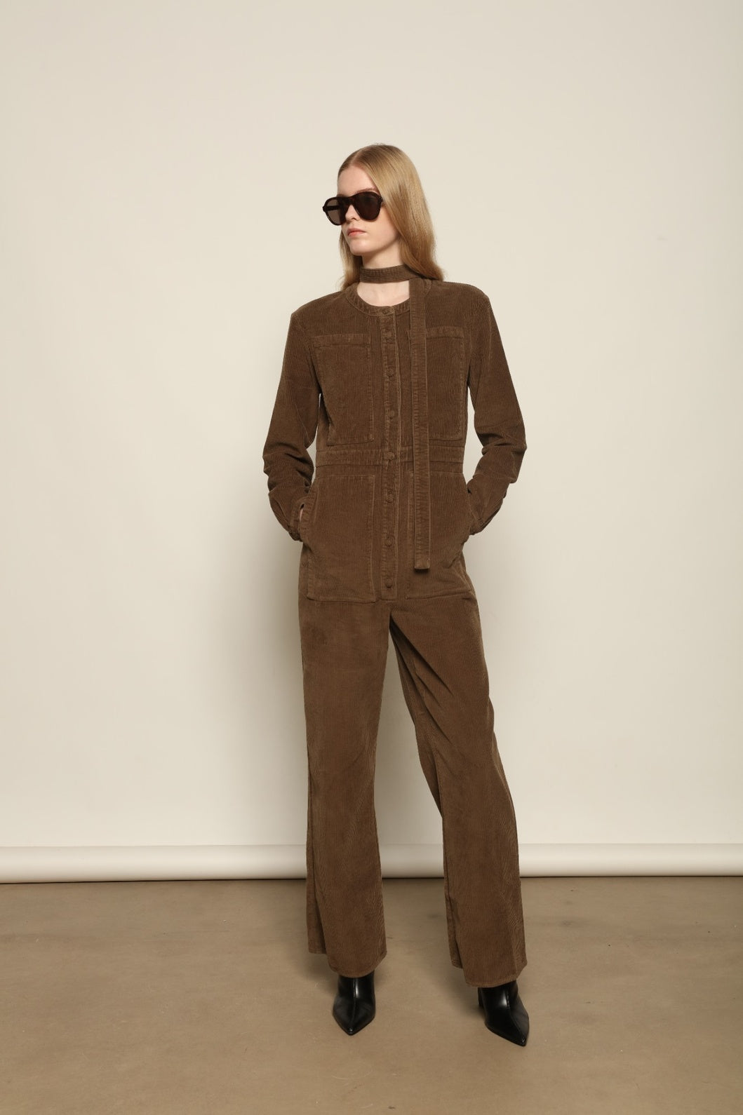 Model is wearing La Combinaison 4 Poches by Happy Haus, a bronze cotton velour jumpsuit with corozo buttons, chest and hip patch pockets, removable belt and relaxed cuffs, curated in the House of Woven Stories selection