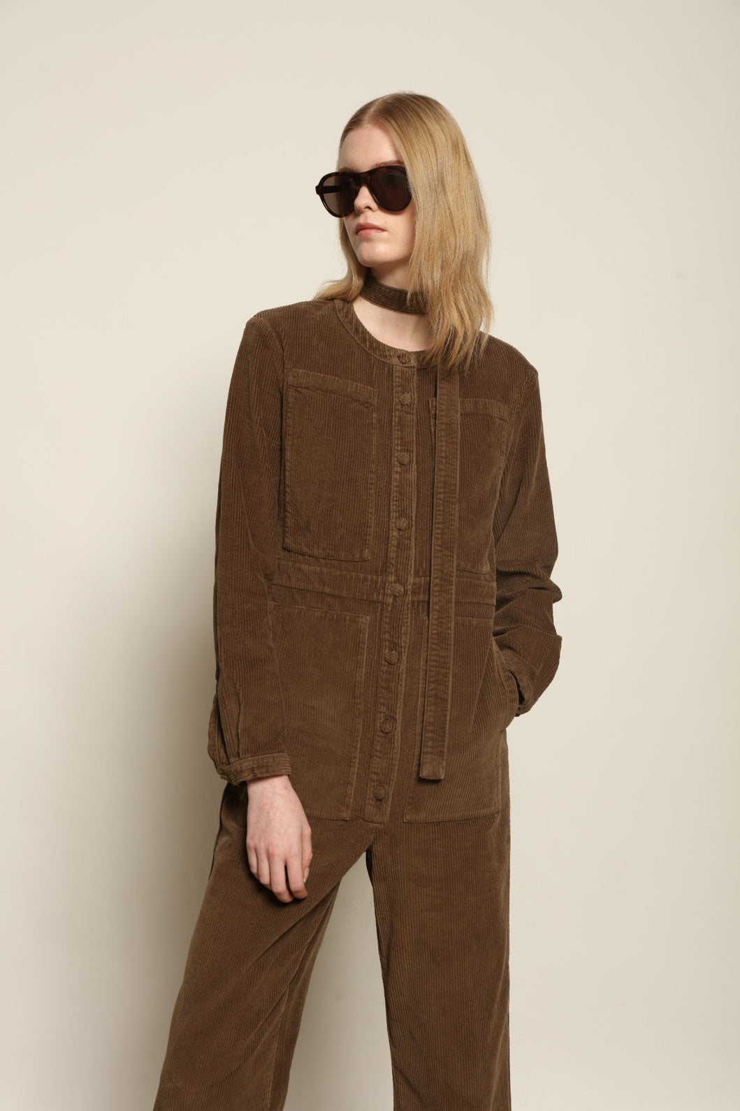 Model is wearing La Combinaison 4 Poches by Happy Haus, a bronze cotton velour jumpsuit with corozo buttons, chest and hip patch pockets, removable belt and relaxed cuffs, curated in the House of Woven Stories selection
