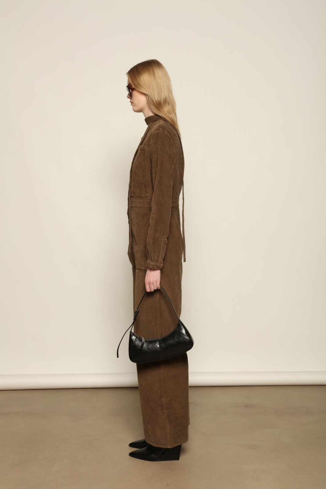 Model is wearing La Combinaison 4 Poches by Happy Haus, a bronze cotton velour jumpsuit with corozo buttons, chest and hip patch pockets, removable belt and relaxed cuffs, curated in the House of Woven Stories selection