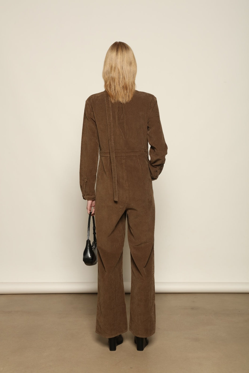 Model is wearing La Combinaison 4 Poches by Happy Haus, a bronze cotton velour jumpsuit with corozo buttons, chest and hip patch pockets, removable belt and relaxed cuffs, curated in the House of Woven Stories selection