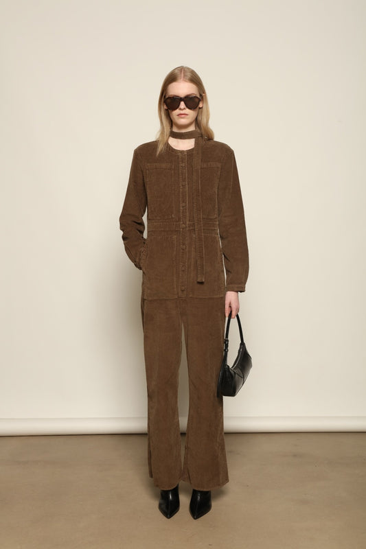 Model is wearing La Combinaison 4 Poches by Happy Haus, a bronze cotton velour jumpsuit with corozo buttons, chest and hip patch pockets, removable belt and relaxed cuffs, curated in the House of Woven Stories selection
