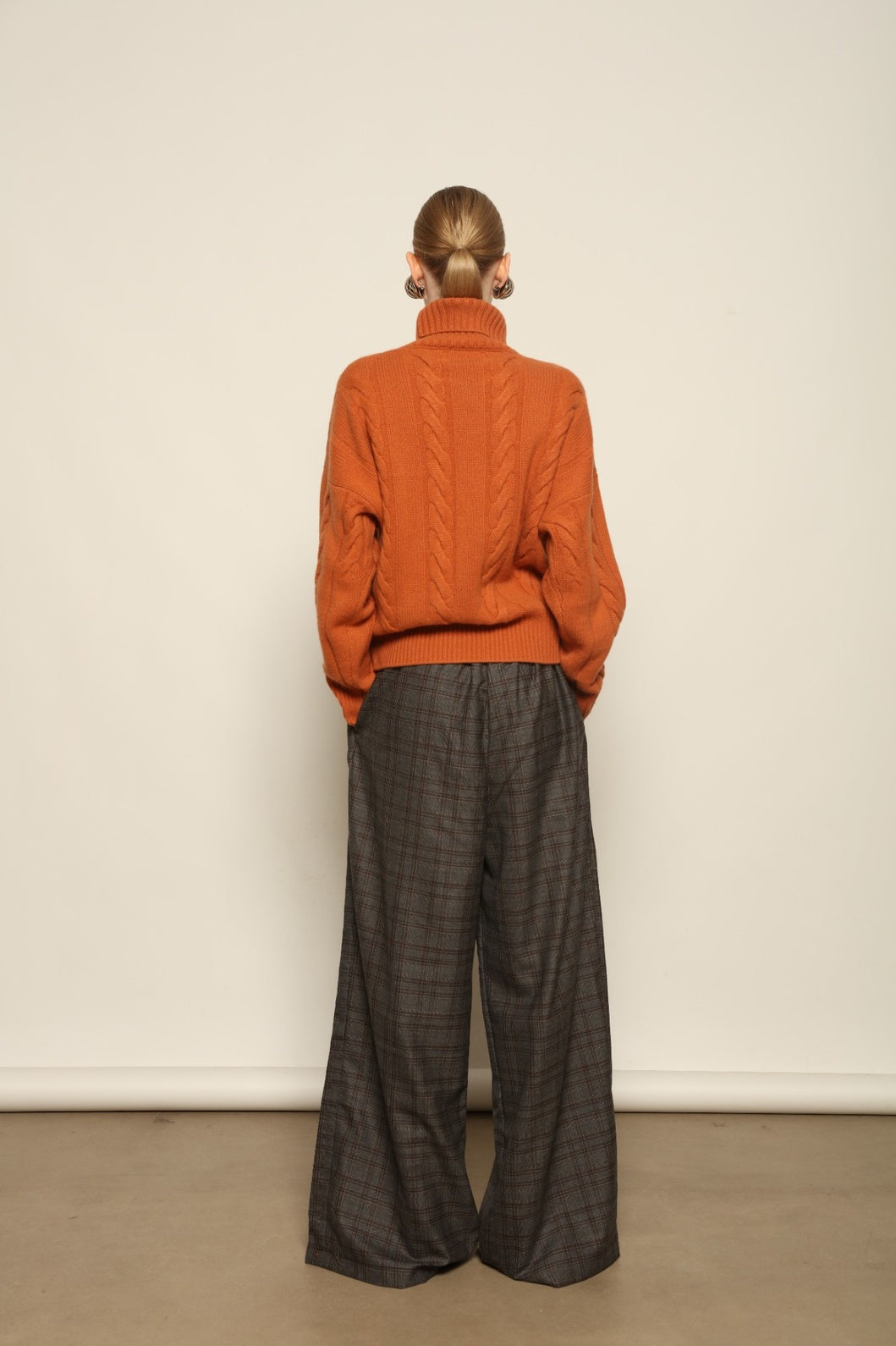 Model is wearing the Brick Noorah Turtleneck Sweater by House of Woven Stories, a pure ethically sourced cashmere cable-knit sweater in a rich brick tone with ribbed high collar and cuffs.