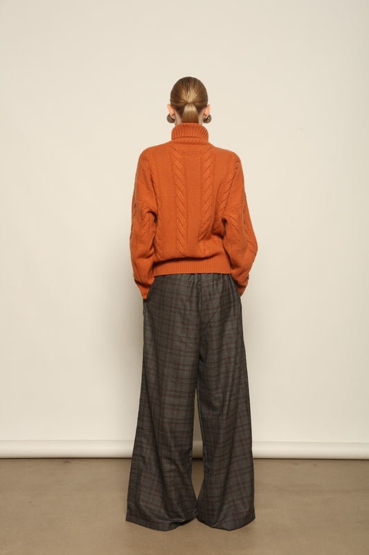 Model is wearing the Brick Noorah Turtleneck Sweater by House of Woven Stories, a pure ethically sourced cashmere cable-knit sweater in a rich brick tone with ribbed high collar and cuffs.