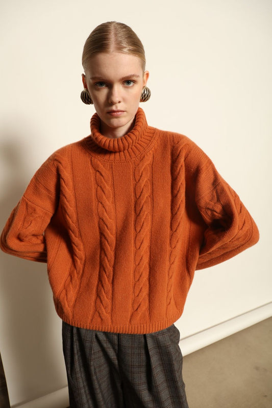 Model is wearing the Brick Noorah Turtleneck Sweater by House of Woven Stories, a pure ethically sourced cashmere cable-knit sweater in a rich brick tone with ribbed high collar and cuffs.