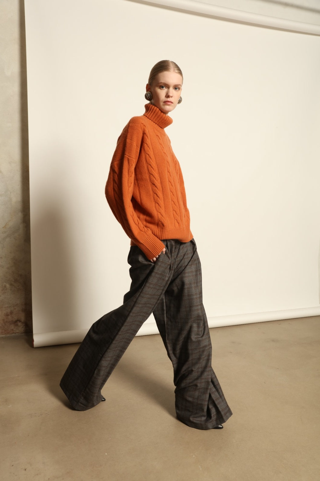 Model is wearing the Brick Noorah Turtleneck Sweater by House of Woven Stories, a pure ethically sourced cashmere cable-knit sweater in a rich brick tone with ribbed high collar and cuffs.