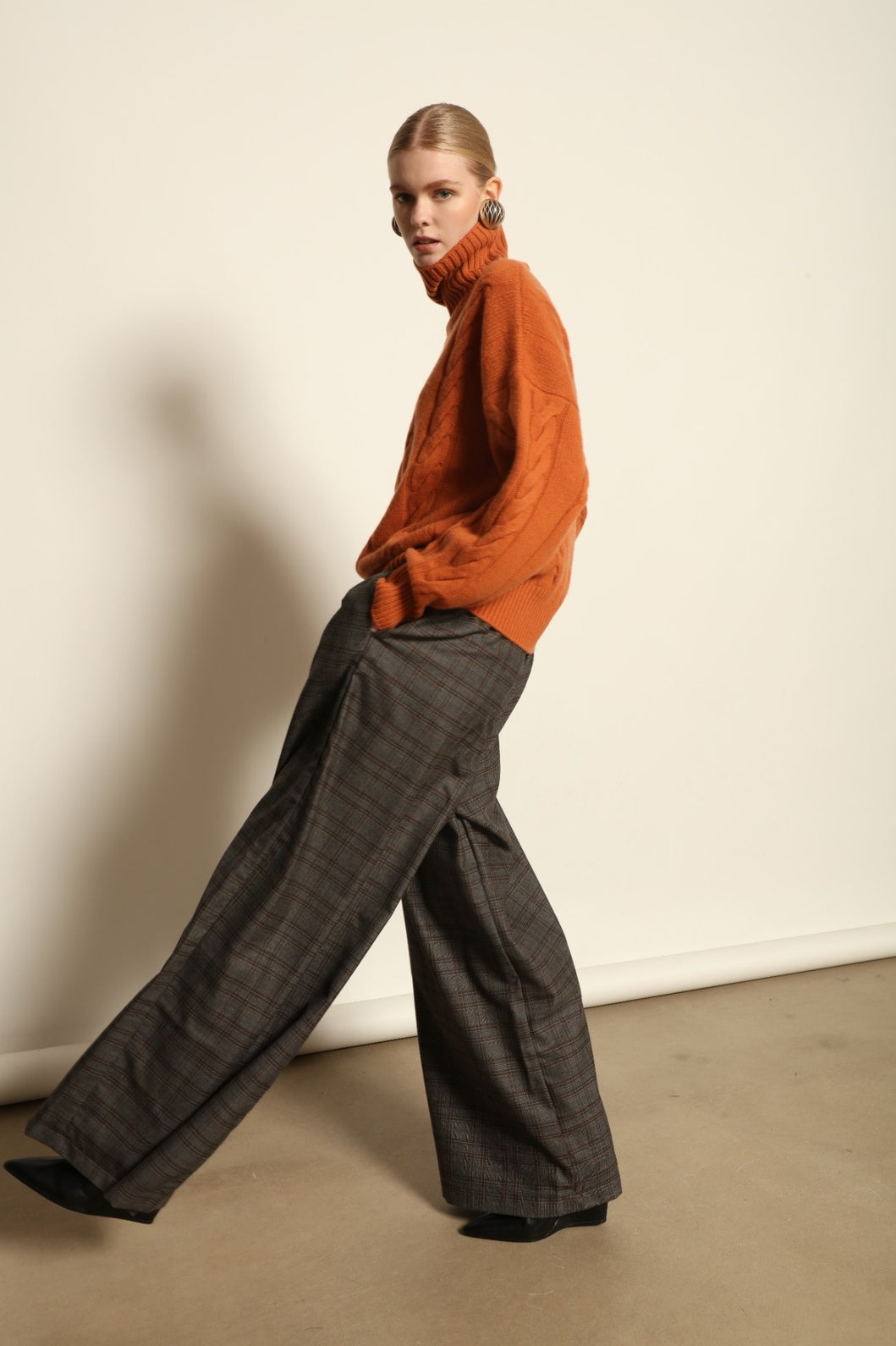 Model is wearing the Brick Noorah Turtleneck Sweater by House of Woven Stories, a pure ethically sourced cashmere cable-knit sweater in a rich brick tone with ribbed high collar and cuffs.