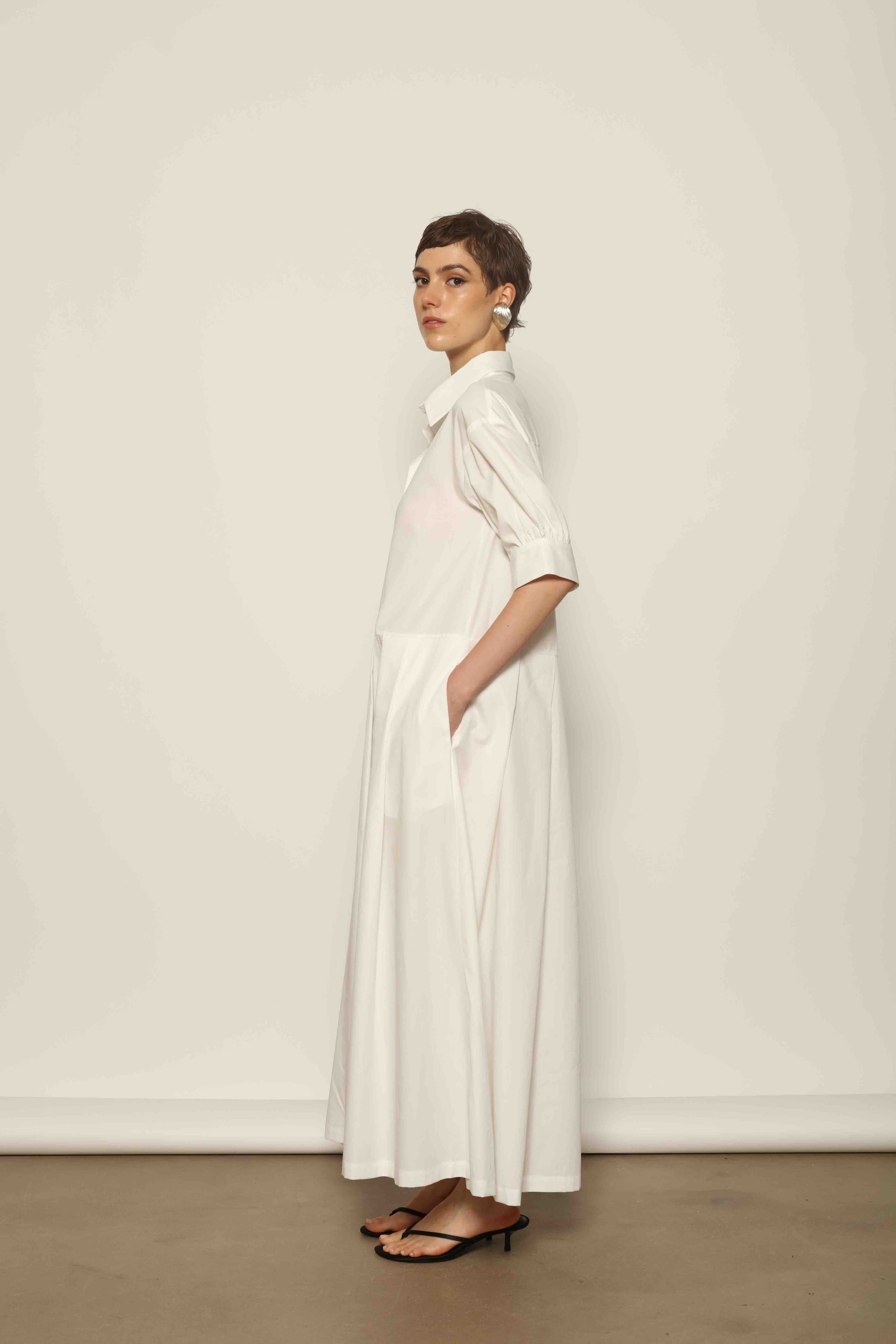 Model wearing the Emily Organic White Cotton Dress by House of Woven Stories, featuring 100% organic cotton fabric, classic collar, button-down front, and adjustable belt for a clean, elegant silhouette.