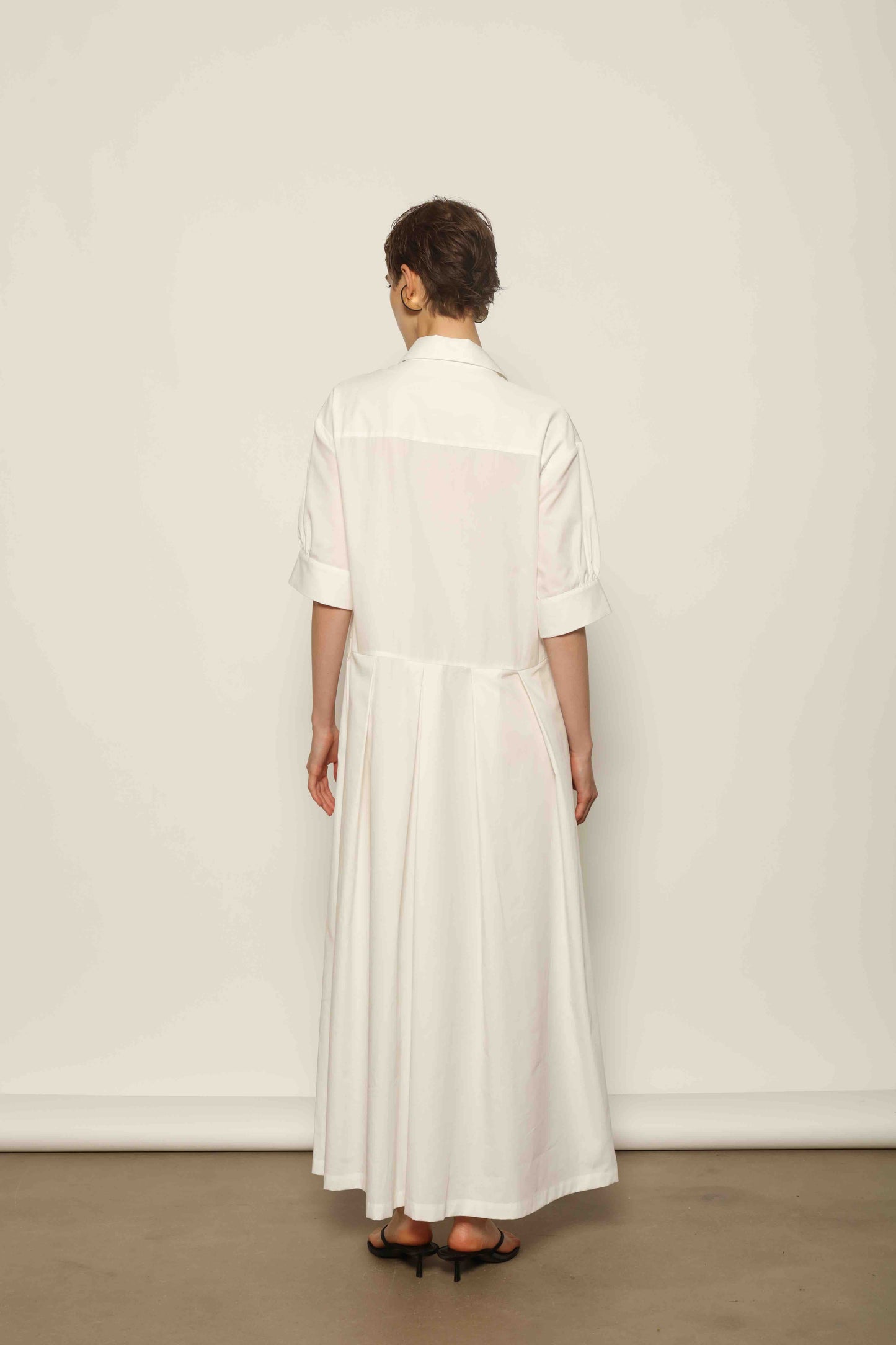 Model wearing the Emily Organic White Cotton Dress by House of Woven Stories, featuring 100% organic cotton fabric, classic collar, button-down front, and adjustable belt for a clean, elegant silhouette.