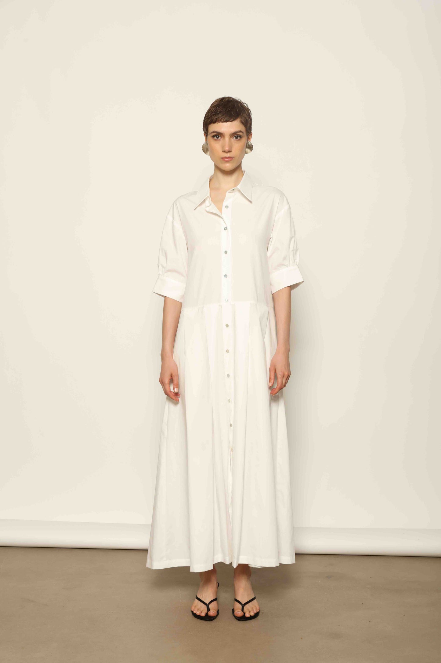 Model wearing the Emily Organic White Cotton Dress by House of Woven Stories, featuring 100% organic cotton fabric, classic collar, button-down front, and adjustable belt for a clean, elegant silhouette.