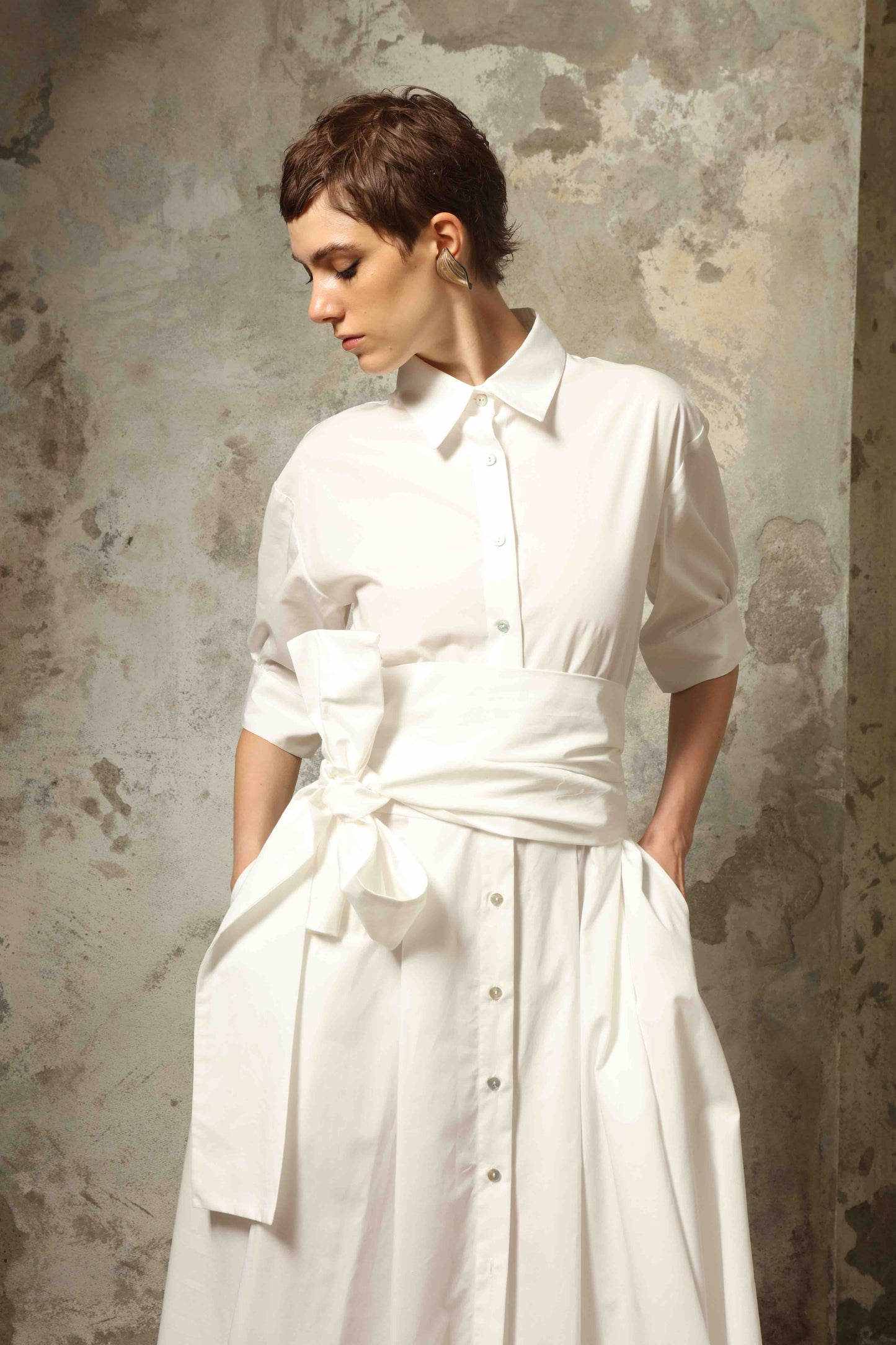 Model wearing the Emily Organic White Cotton Dress by House of Woven Stories, featuring 100% organic cotton fabric, classic collar, button-down front, and adjustable belt for a clean, elegant silhouette.