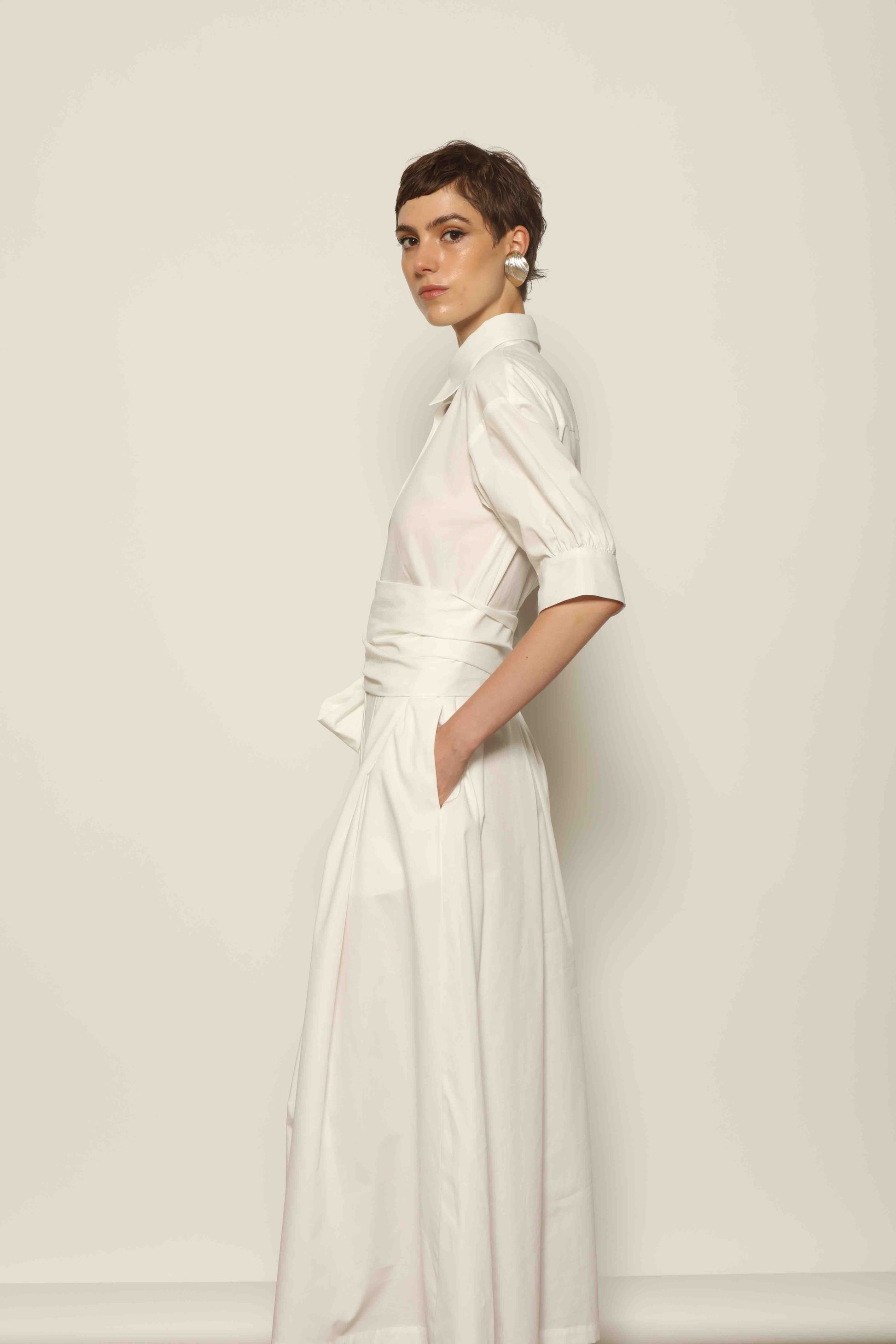 Model wearing the Emily Organic White Cotton Dress by House of Woven Stories, featuring 100% organic cotton fabric, classic collar, button-down front, and adjustable belt for a clean, elegant silhouette.