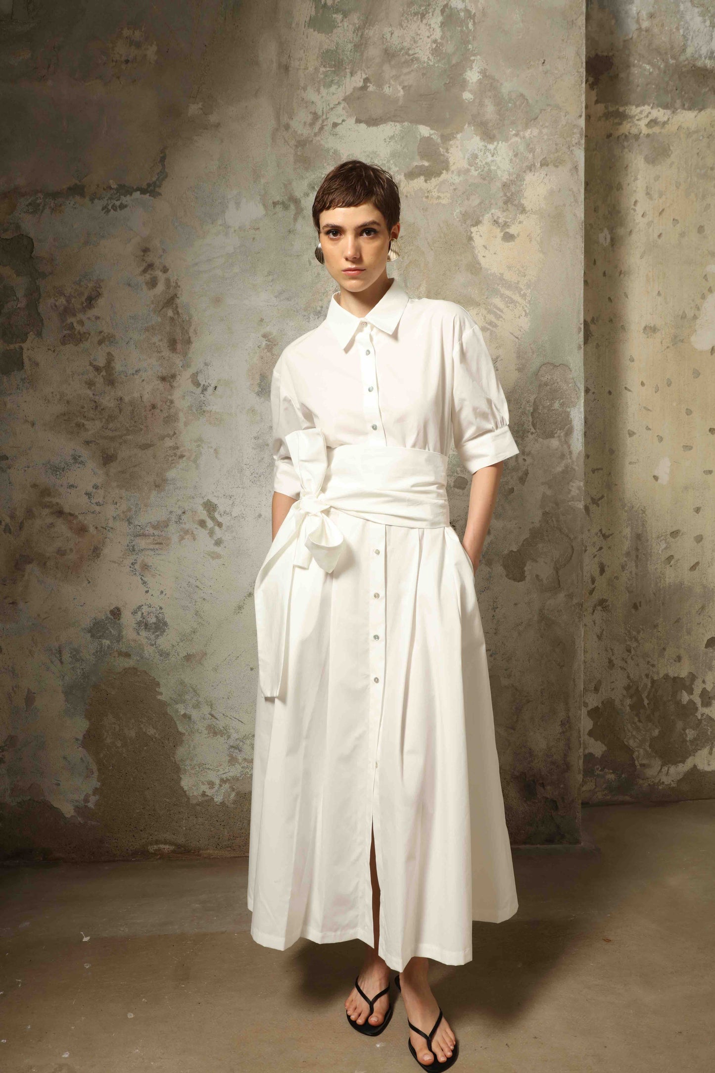 Model wearing the Emily Organic White Cotton Dress by House of Woven Stories, featuring 100% organic cotton fabric, classic collar, button-down front, and adjustable belt for a clean, elegant silhouette.