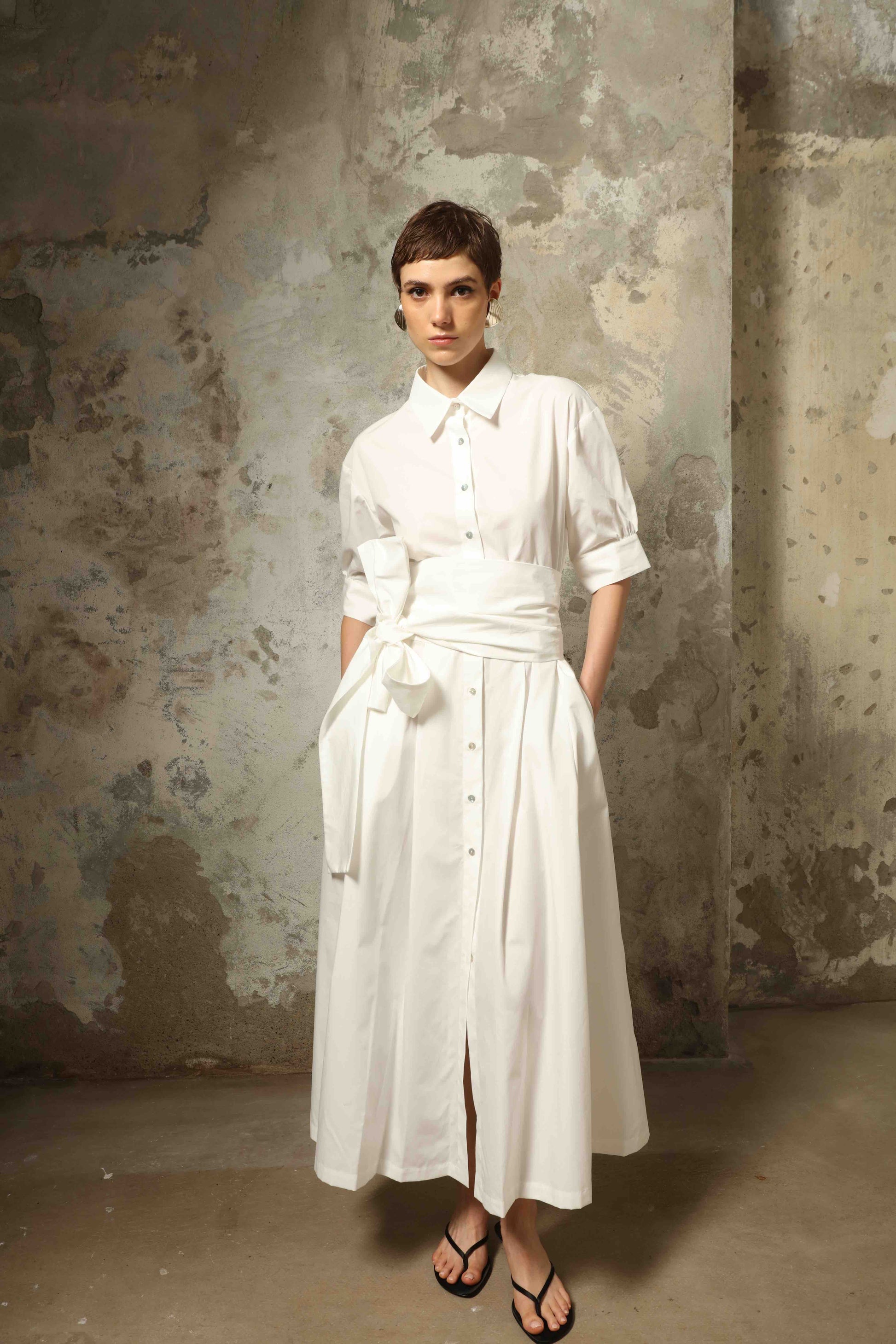 Model wearing the Emily Organic White Cotton Dress by House of Woven Stories, featuring 100% organic cotton fabric, classic collar, button-down front, and adjustable belt for a clean, elegant silhouette.