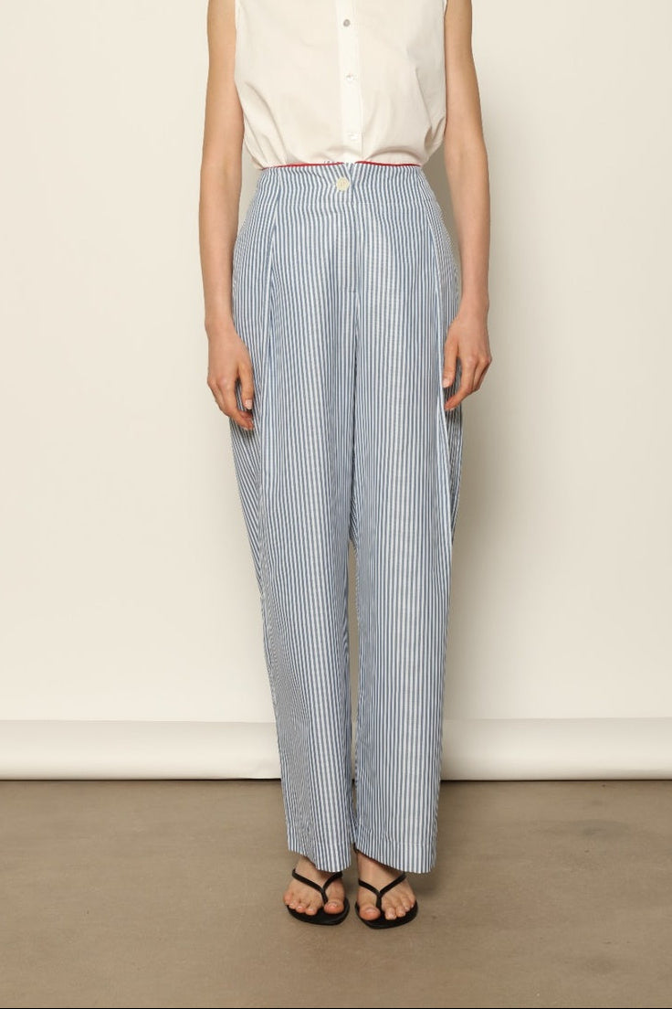Model wearing the Gabriella Blue & White Striped Trousers by House of Woven Stories, featuring 100% cotton fabric, blue and white stripe pattern, relaxed fit, and contrasting red piping at the waistband.