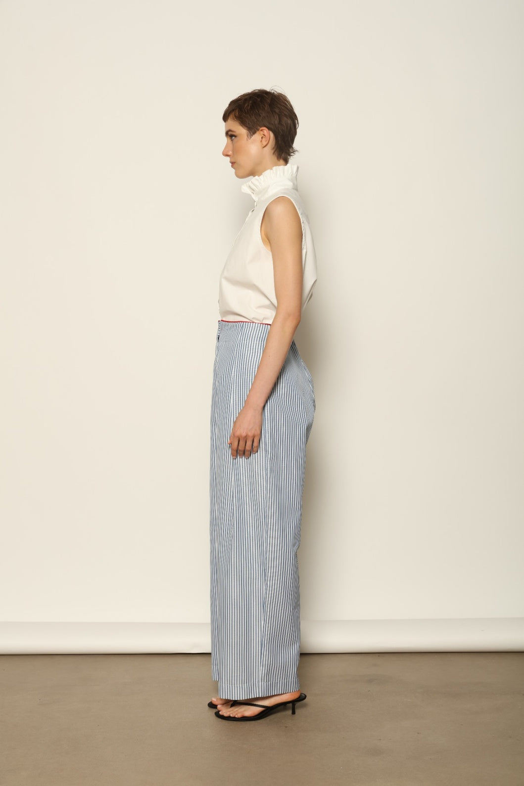 Model wearing the Gabriella Blue & White Striped Trousers by House of Woven Stories, featuring 100% cotton fabric, blue and white stripe pattern, relaxed fit, and contrasting red piping at the waistband.