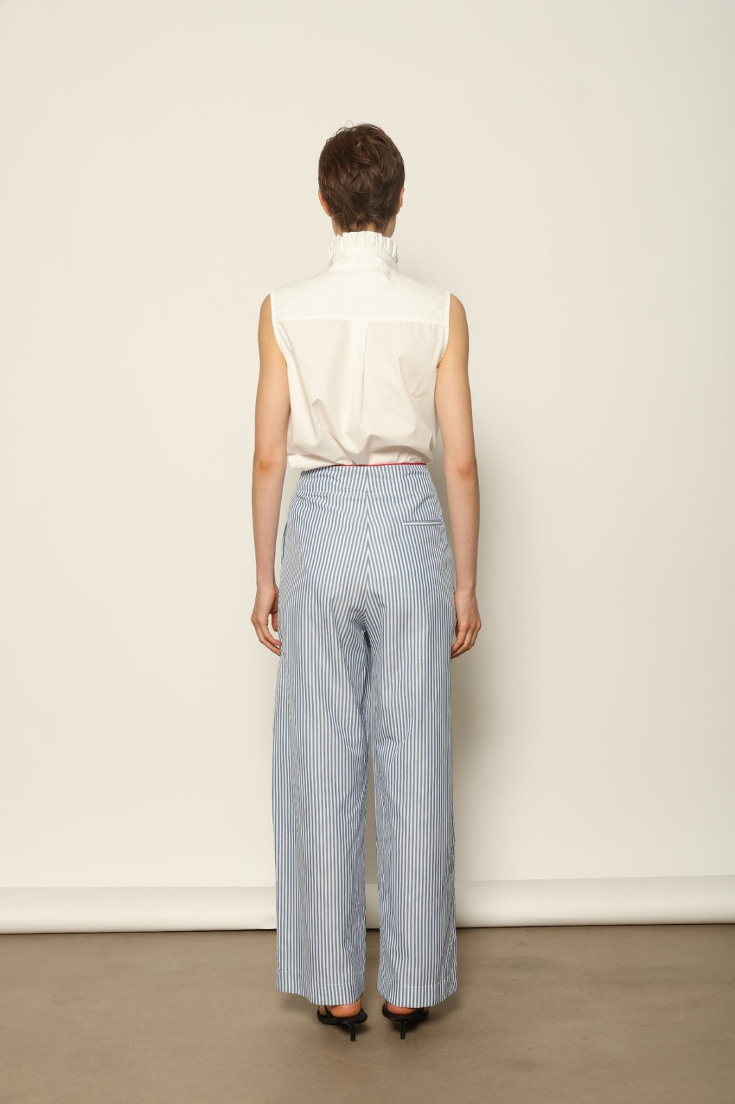 Model wearing the Gabriella Blue & White Striped Trousers by House of Woven Stories, featuring 100% cotton fabric, blue and white stripe pattern, relaxed fit, and contrasting red piping at the waistband.