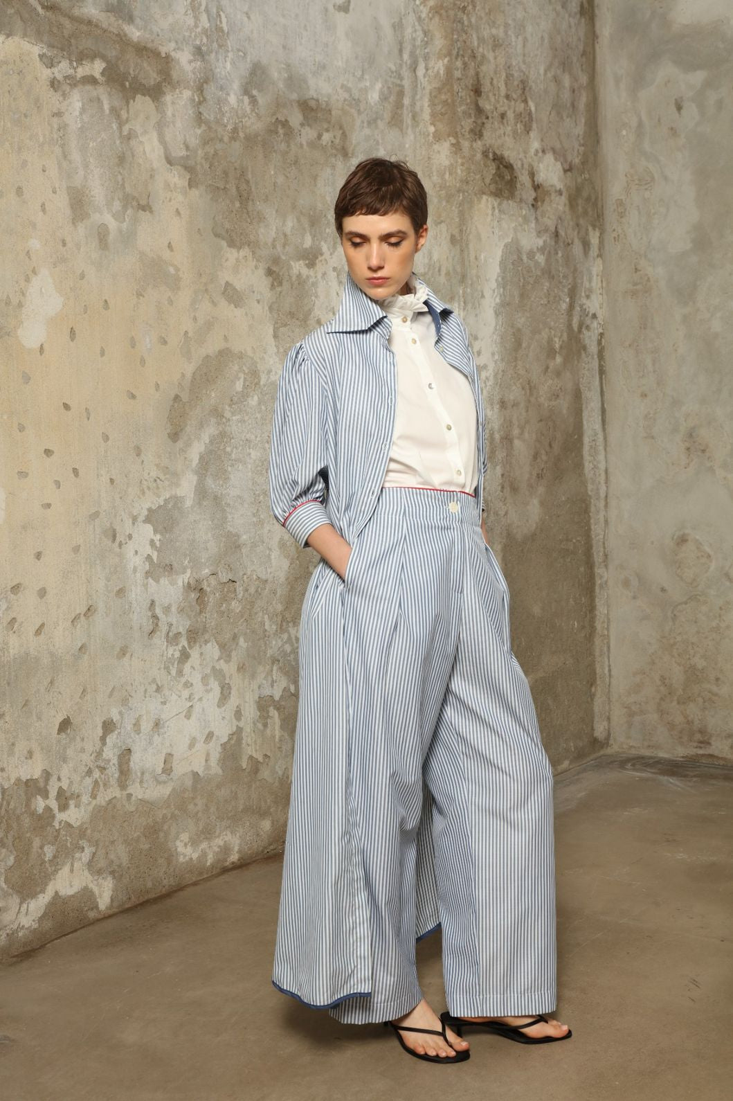 Model wearing the Gabriella Blue & White Striped Trousers by House of Woven Stories, featuring 100% cotton fabric, blue and white stripe pattern, relaxed fit, and contrasting red piping at the waistband.