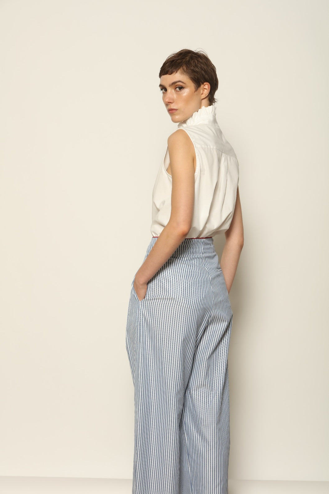 Model wearing the Gabriella Blue & White Striped Trousers by House of Woven Stories, featuring 100% cotton fabric, blue and white stripe pattern, relaxed fit, and contrasting red piping at the waistband.