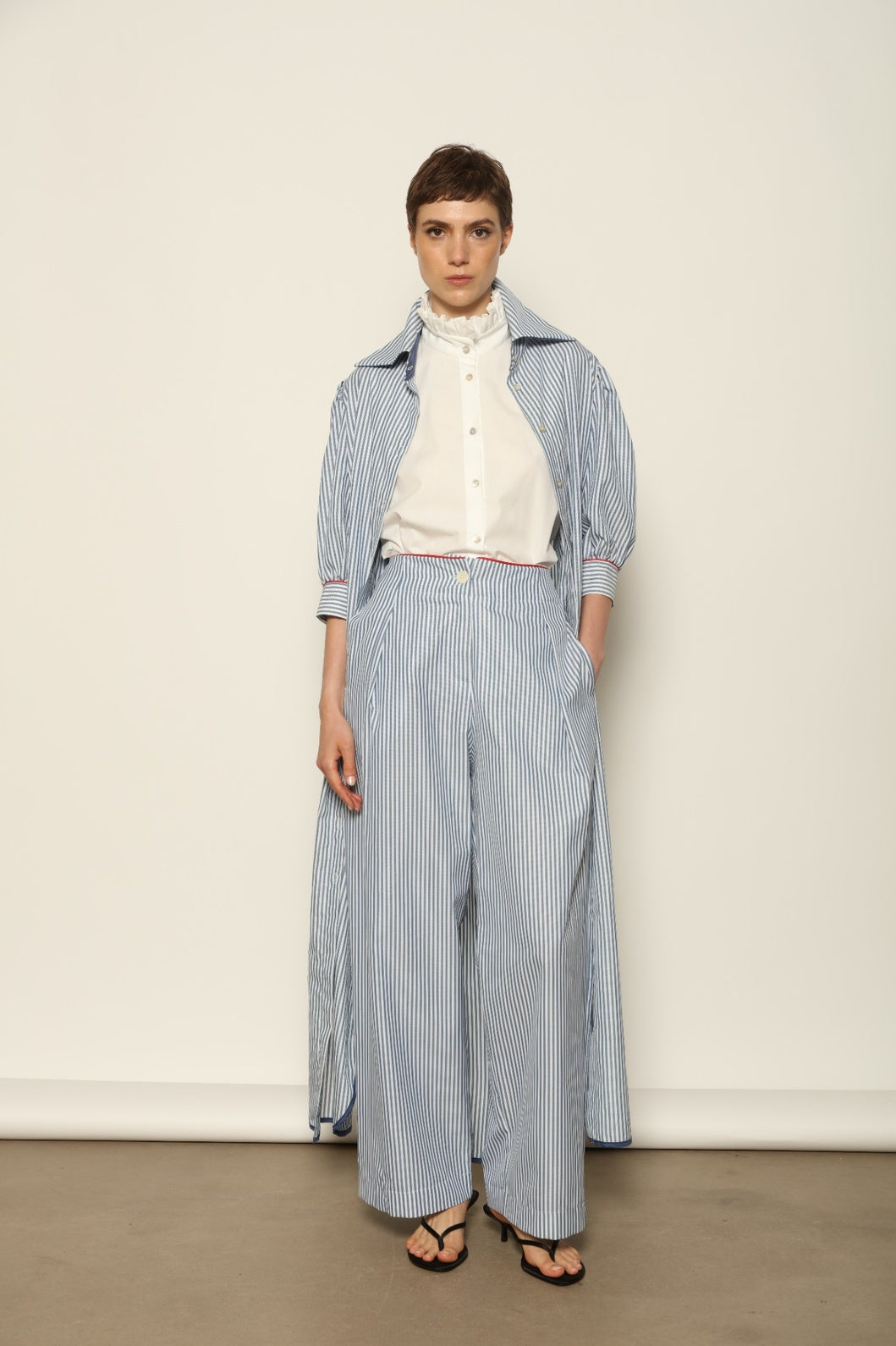 Model wearing the Gabriella Blue & White Striped Trousers by House of Woven Stories, featuring 100% cotton fabric, blue and white stripe pattern, relaxed fit, and contrasting red piping at the waistband.