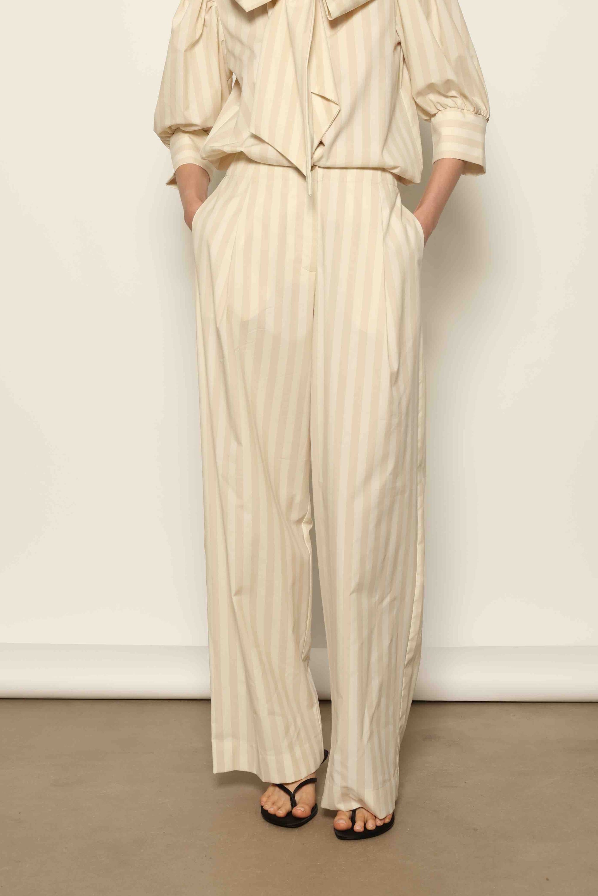Model wearing the Gabriella Sand White Striped Trousers by House of Woven Stories, featuring a high-rise waist, straight-leg tailored fit, and soft sand and white stripe pattern in a premium cotton, merino wool, and cashmere blend.