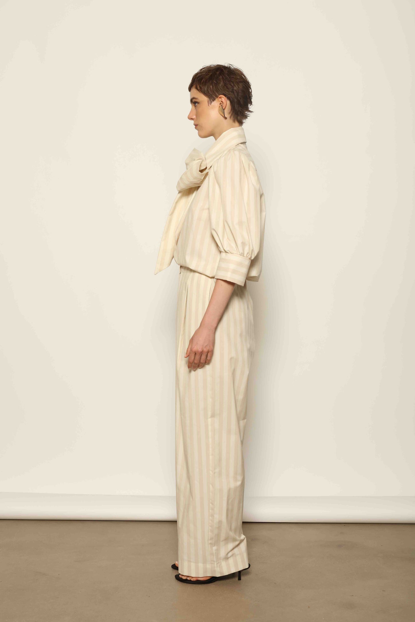 Model wearing the Gabriella Sand White Striped Trousers by House of Woven Stories, featuring a high-rise waist, straight-leg tailored fit, and soft sand and white stripe pattern in a premium cotton, merino wool, and cashmere blend.