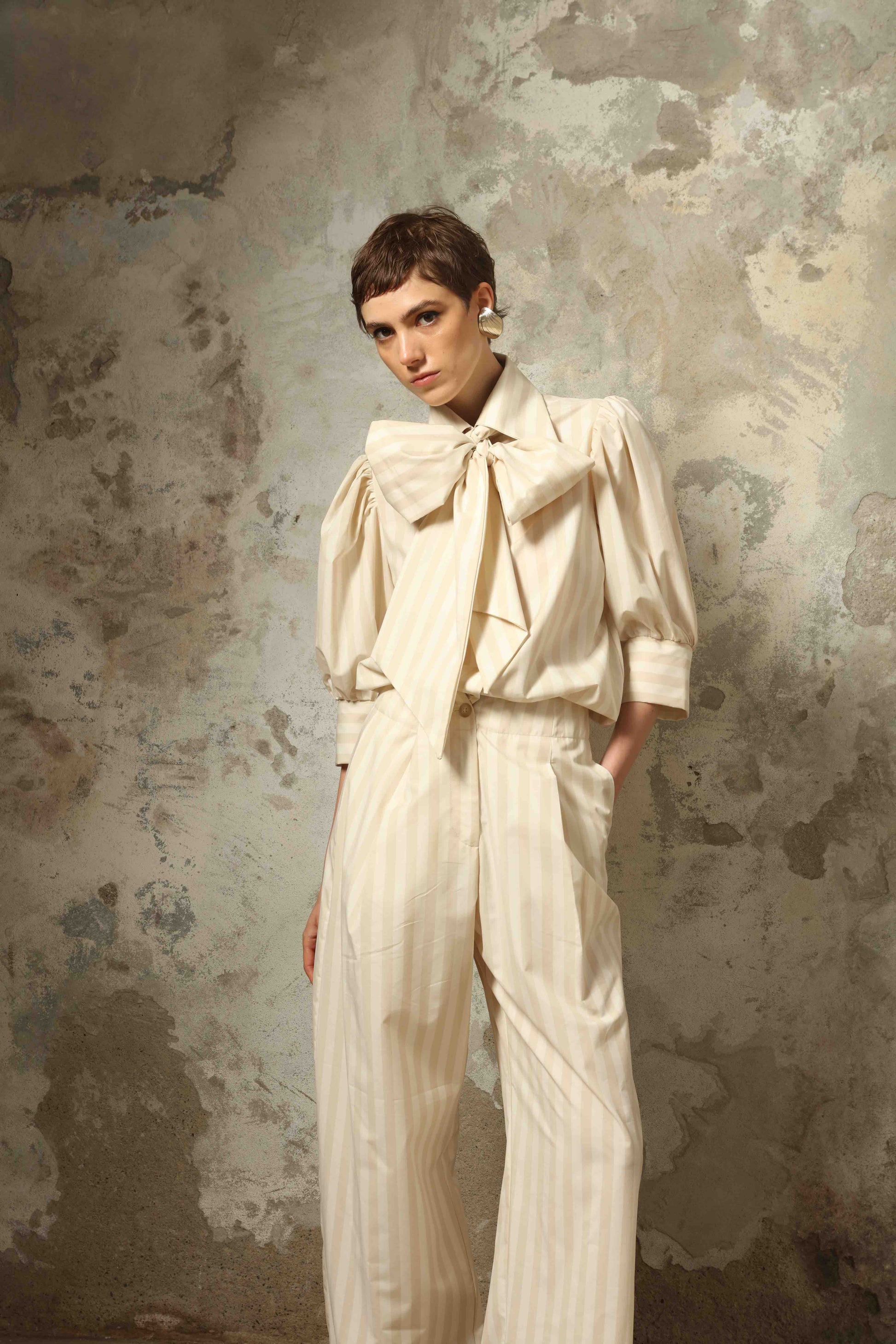 Model wearing the Gabriella Sand White Striped Trousers by House of Woven Stories, featuring a high-rise waist, straight-leg tailored fit, and soft sand and white stripe pattern in a premium cotton, merino wool, and cashmere blend.