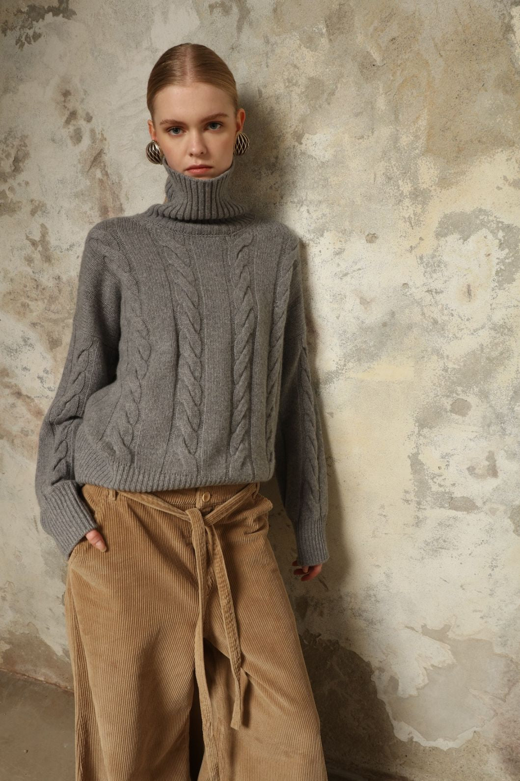 Model is wearing the Graphite Noorah Turtleneck Sweater by House of Woven Stories, a pure ethically sourced cashmere cable-knit sweater with wide ribbed turtleneck and cuffs in graphite.