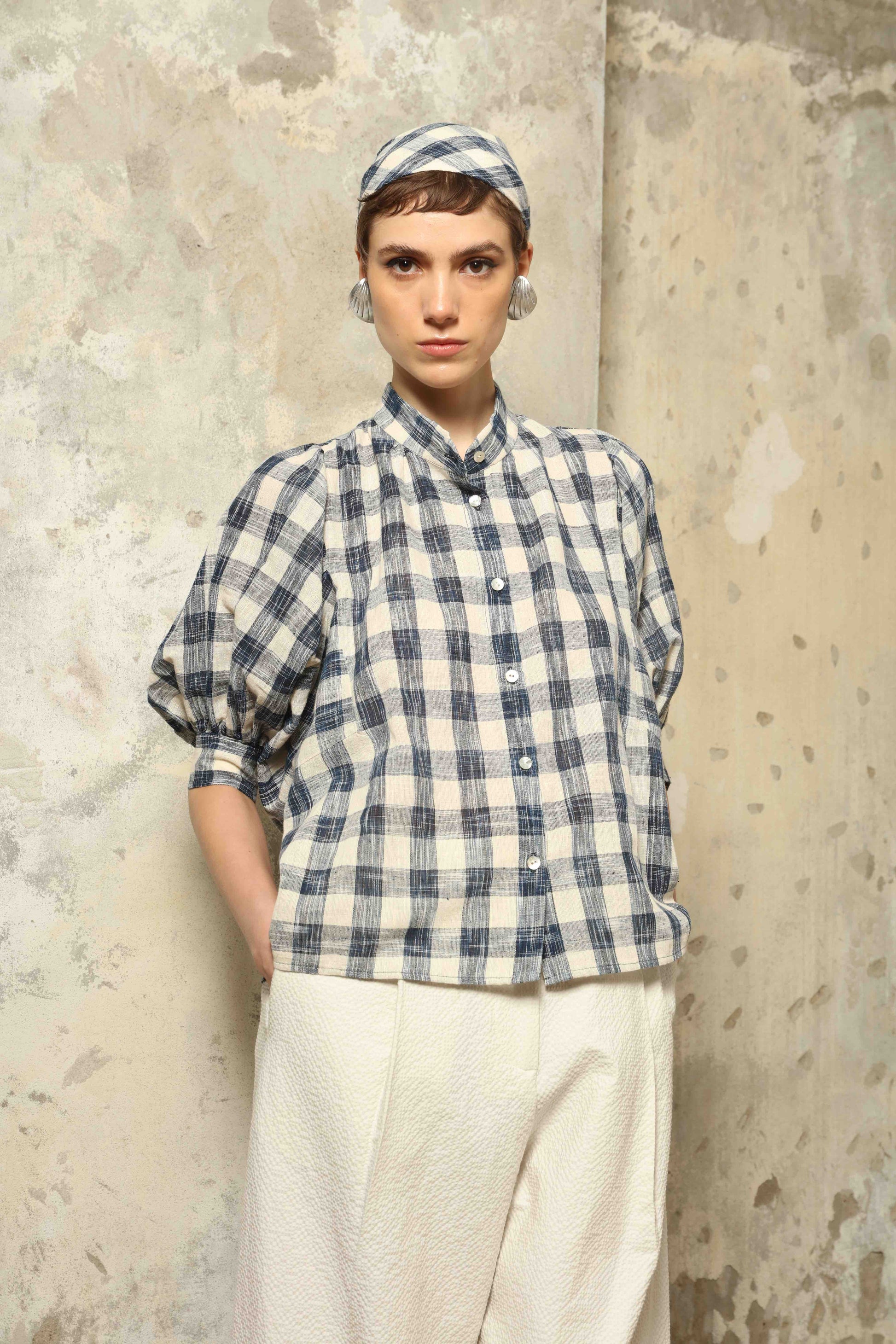 Model wearing the Lola Check Blouse by House of Woven Stories, crafted from 100% handloomed Kala cotton with a blue and white check pattern, Mandarin collar, button-front closure, and balloon sleeves.