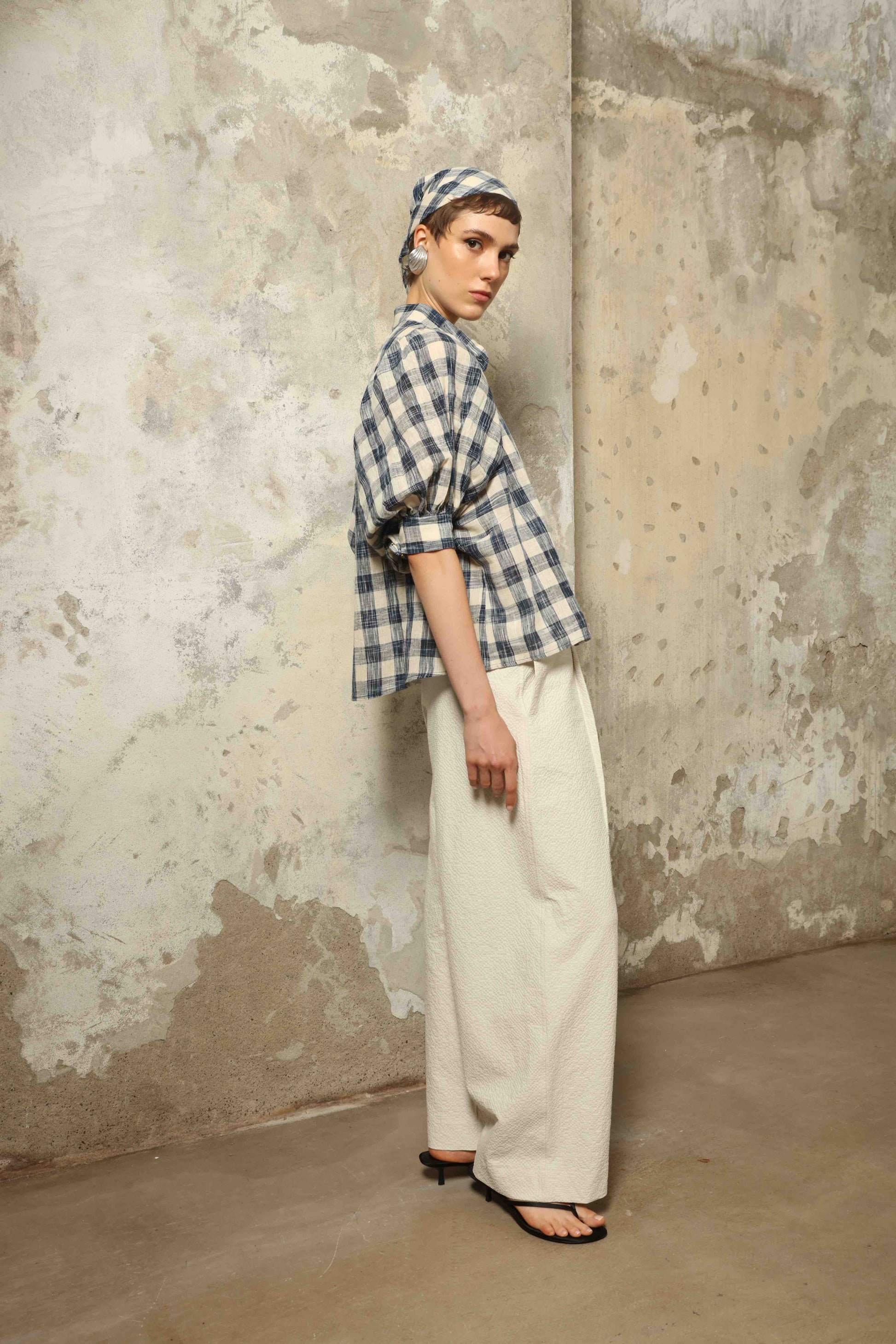 Model wearing the Lola Check Blouse by House of Woven Stories, crafted from 100% handloomed Kala cotton with a blue and white check pattern, Mandarin collar, button-front closure, and balloon sleeves.