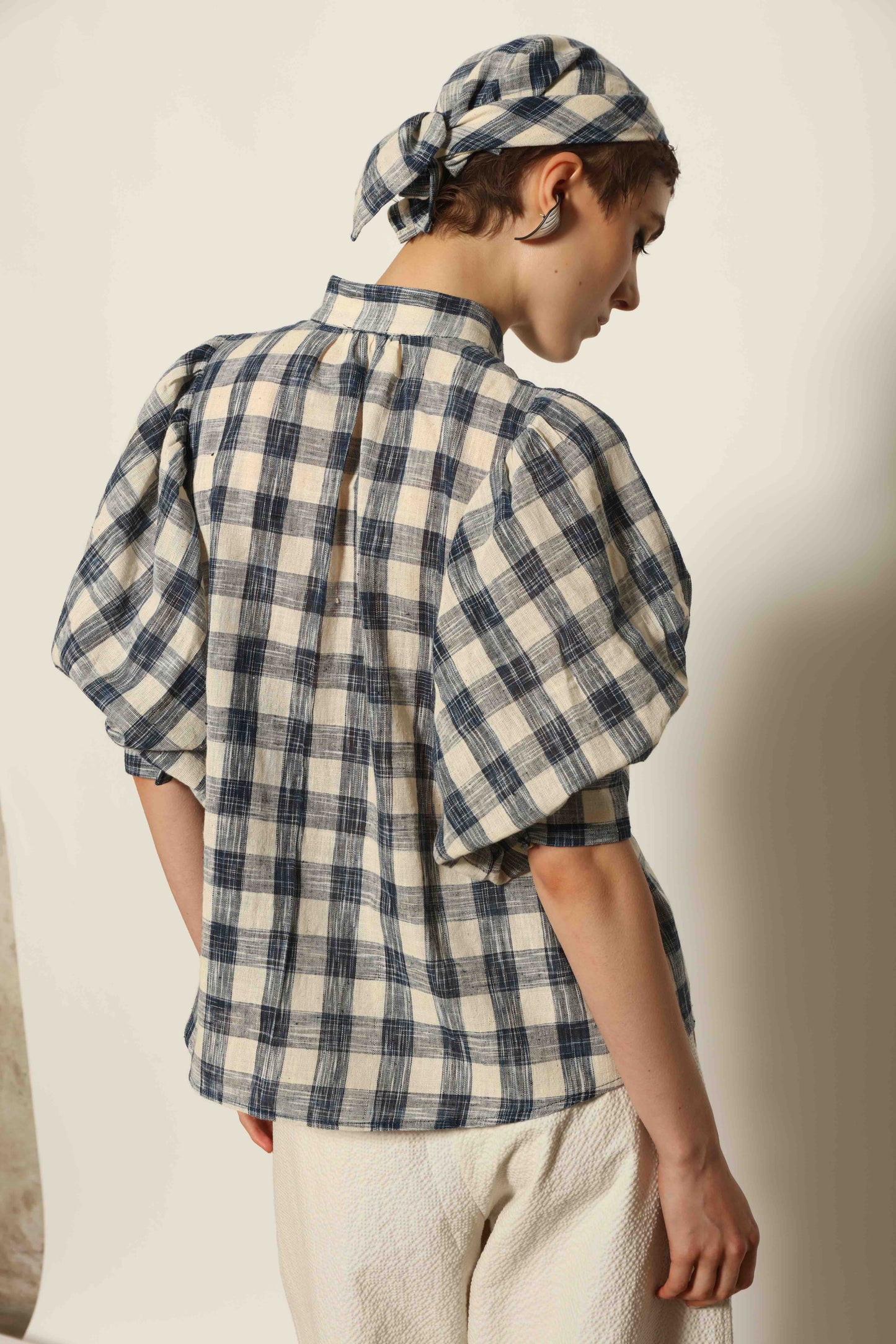 Model wearing the Lola Check Blouse by House of Woven Stories, crafted from 100% handloomed Kala cotton with a blue and white check pattern, Mandarin collar, button-front closure, and balloon sleeves.