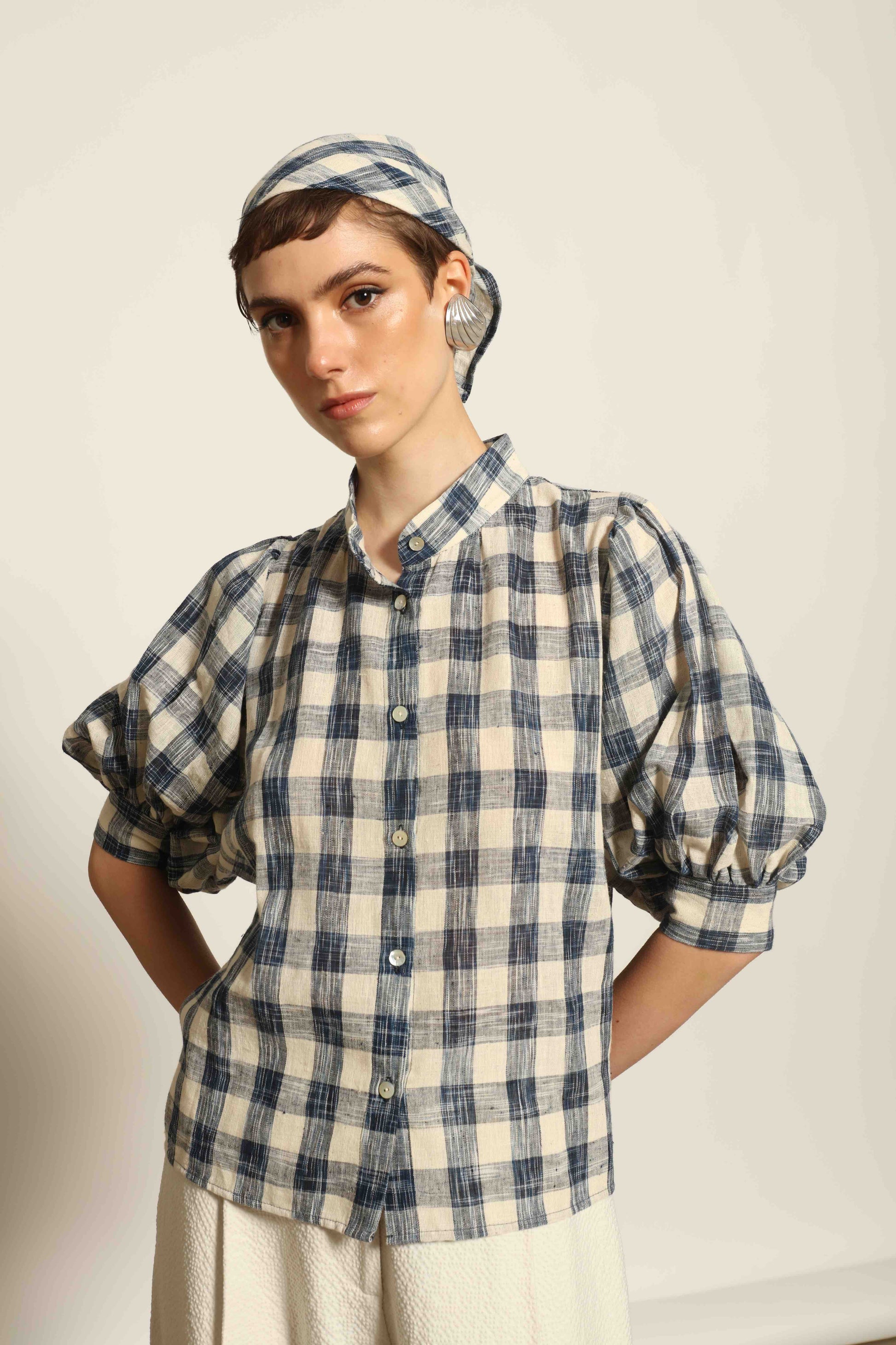 Model wearing the Lola Check Blouse by House of Woven Stories, crafted from 100% handloomed Kala cotton with a blue and white check pattern, Mandarin collar, button-front closure, and balloon sleeves.