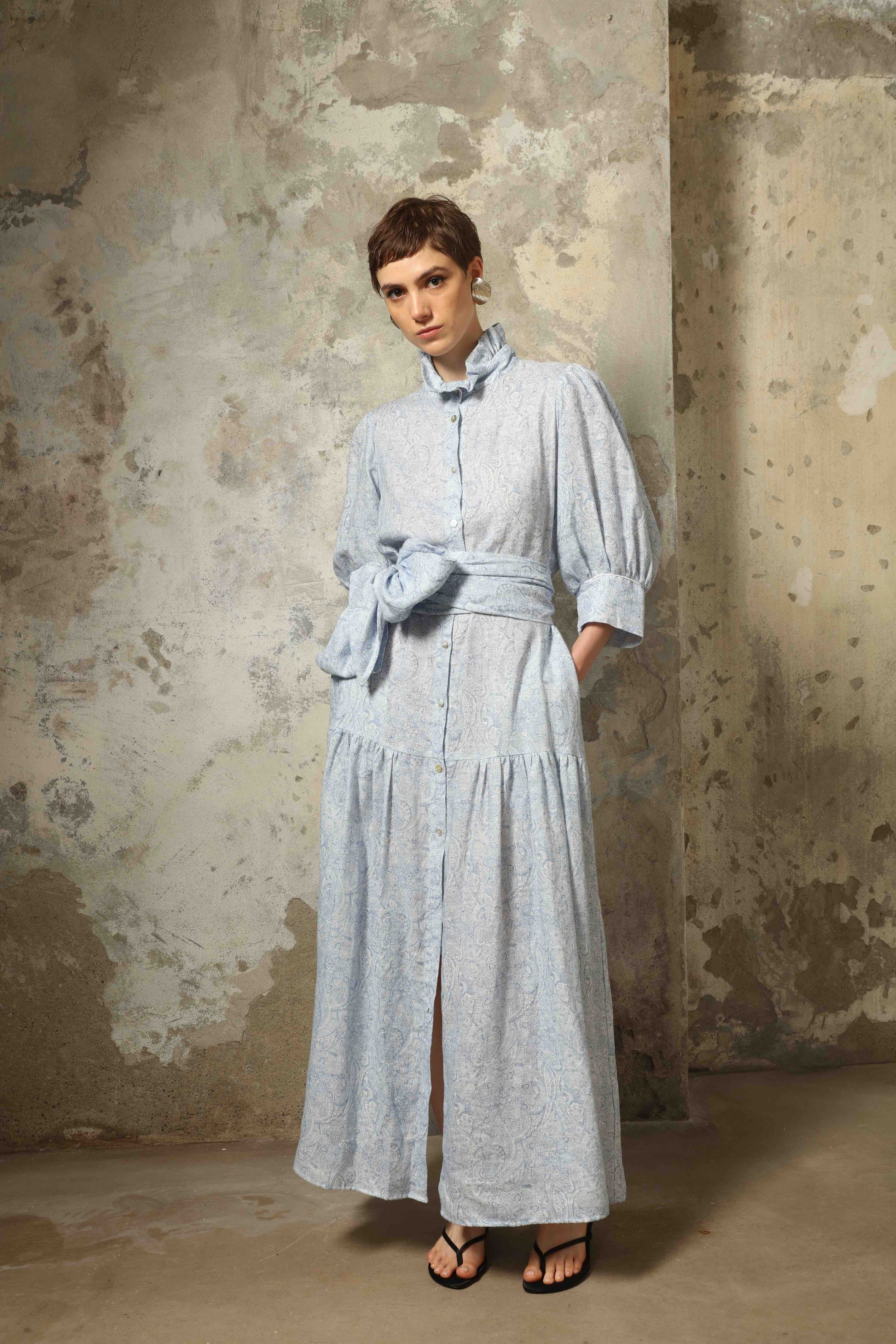 Model wearing the Karin Blue Paisley Dress by House of Woven Stories, featuring 100% linen fabric, soft blue paisley print, and a relaxed flowing silhouette with breathable lightweight structure.